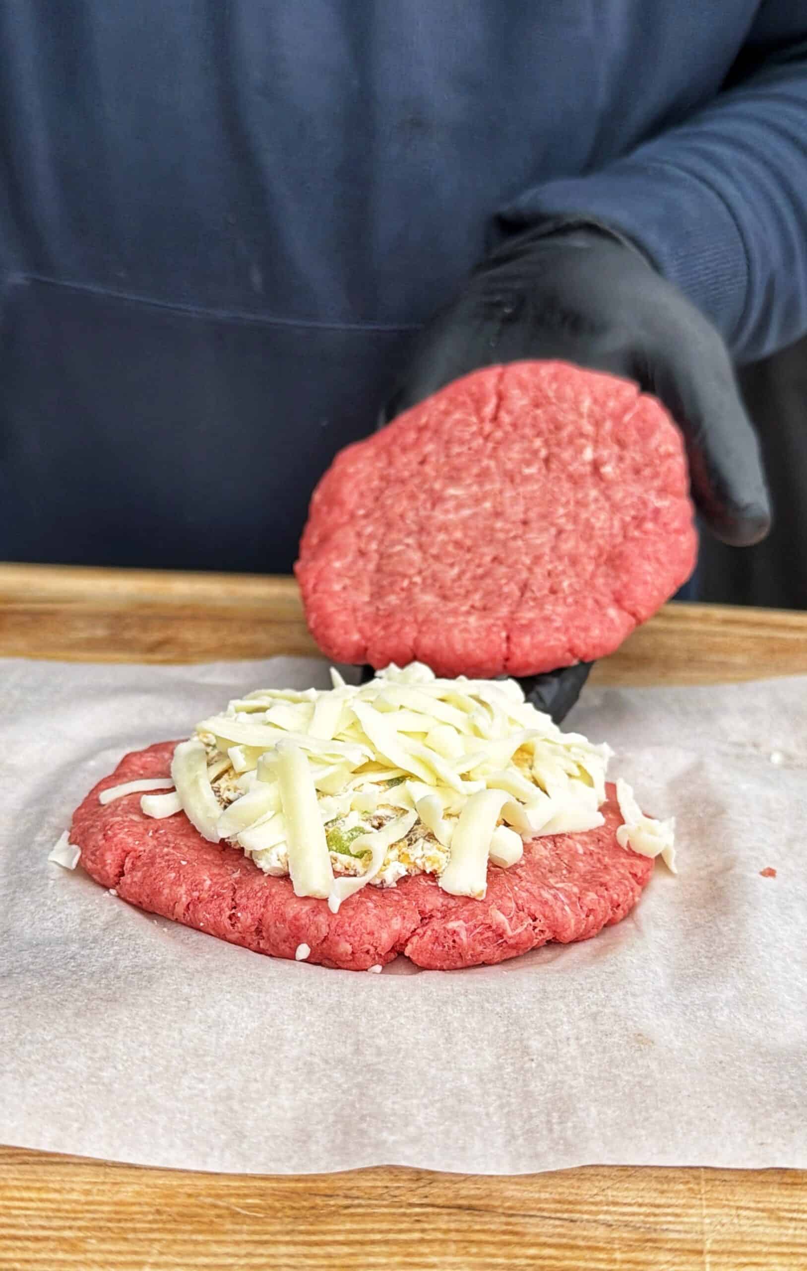 A person wearing black gloves places a raw beef patty on top of another patty with shredded cheese and onions, preparing a stuffed burger on parchment paper.