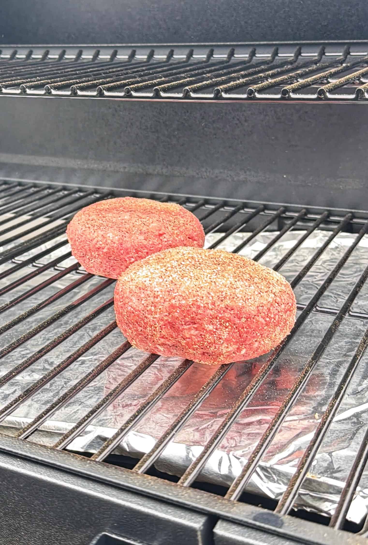 Two seasoned, uncooked burger patties are placed on the lower rack of a grill lined with aluminum foil, ready for cooking. The upper grill rack is empty.