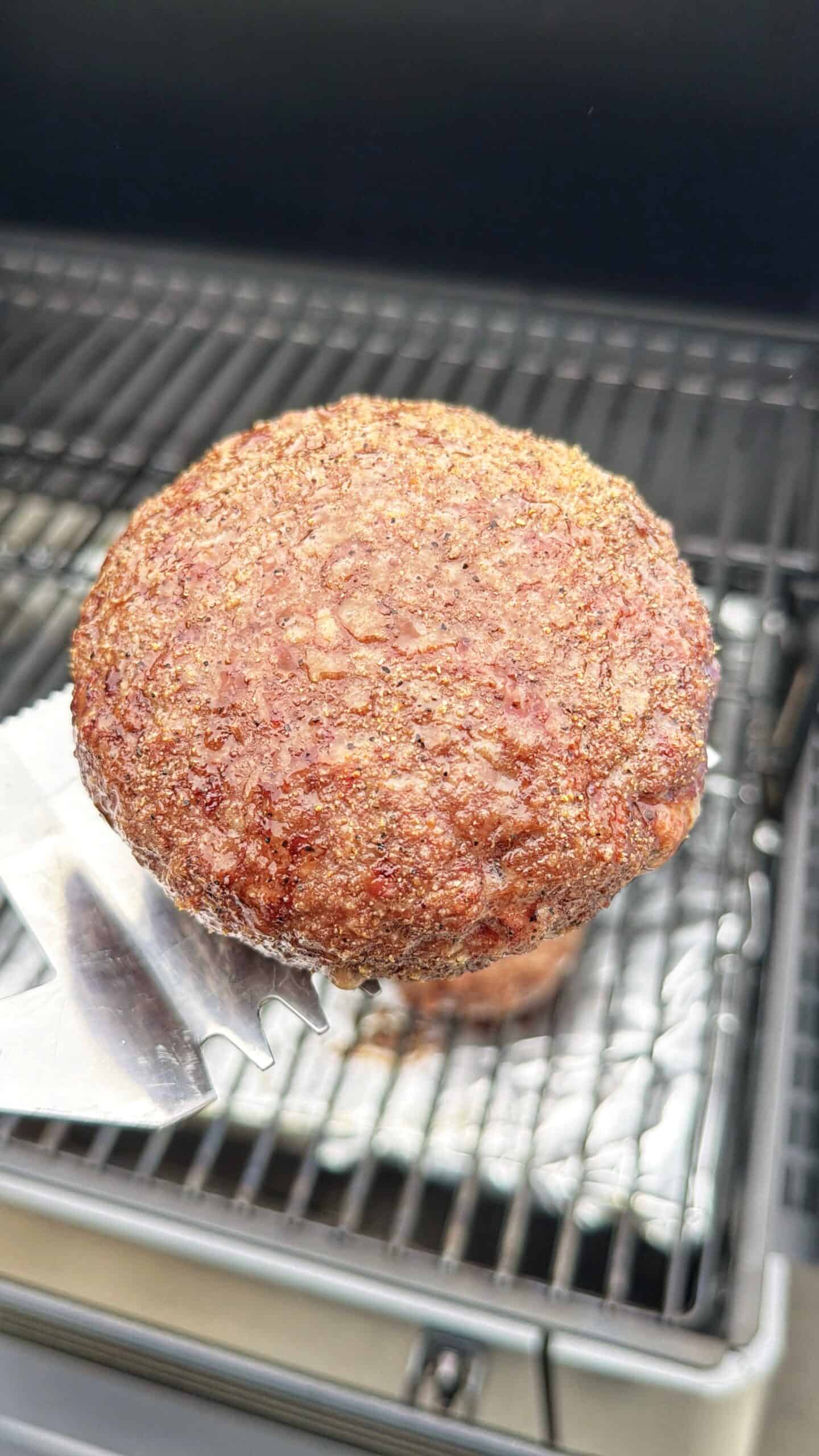 A close-up of a thick, juicy hamburger patty being lifted with a metal spatula from a grill, with grill grates and a foil tray visible in the background.