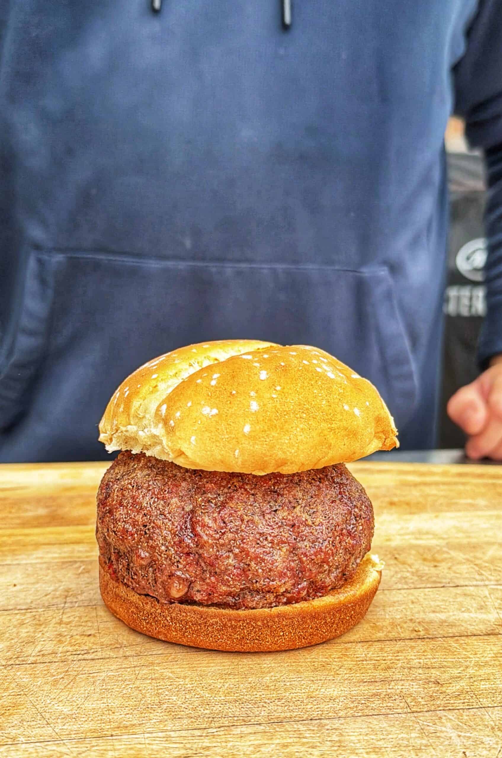 A thick, round beef patty in a sesame seed bun sits on a wooden surface, with a person in a blue hoodie standing in the background.