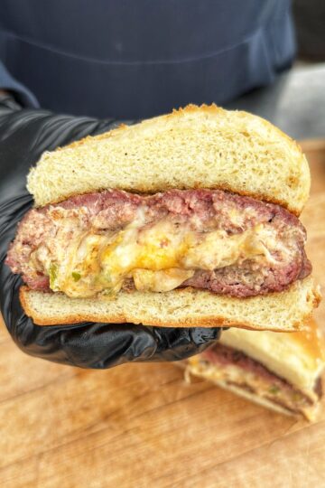 A gloved hand holds a sliced burger showing a thick beef patty stuffed with melted cheese and peppers, between two halves of a soft bun, with another sandwich half on a wooden surface below.