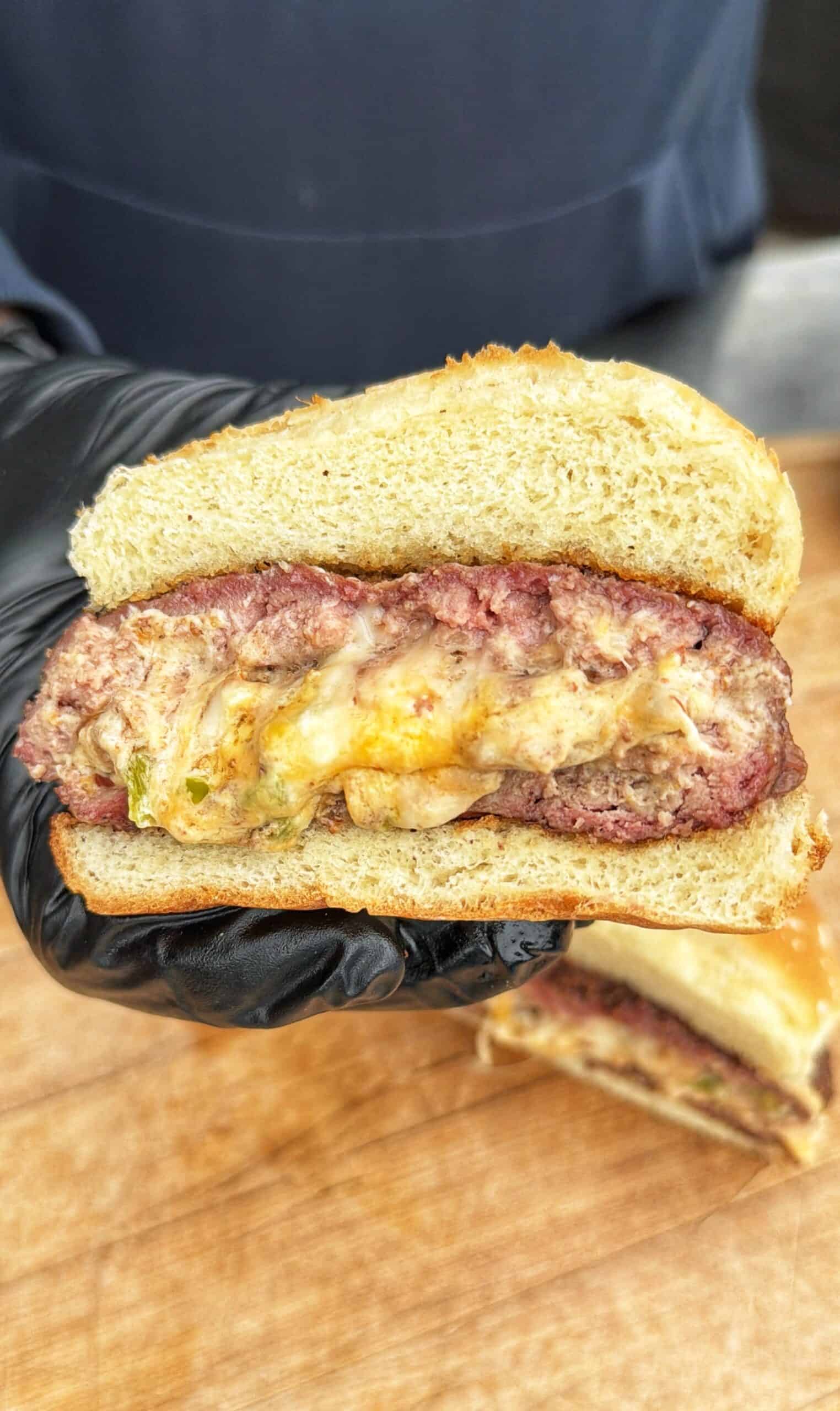A gloved hand holds a sliced burger showing a thick beef patty stuffed with melted cheese and peppers, between two halves of a soft bun, with another sandwich half on a wooden surface below.