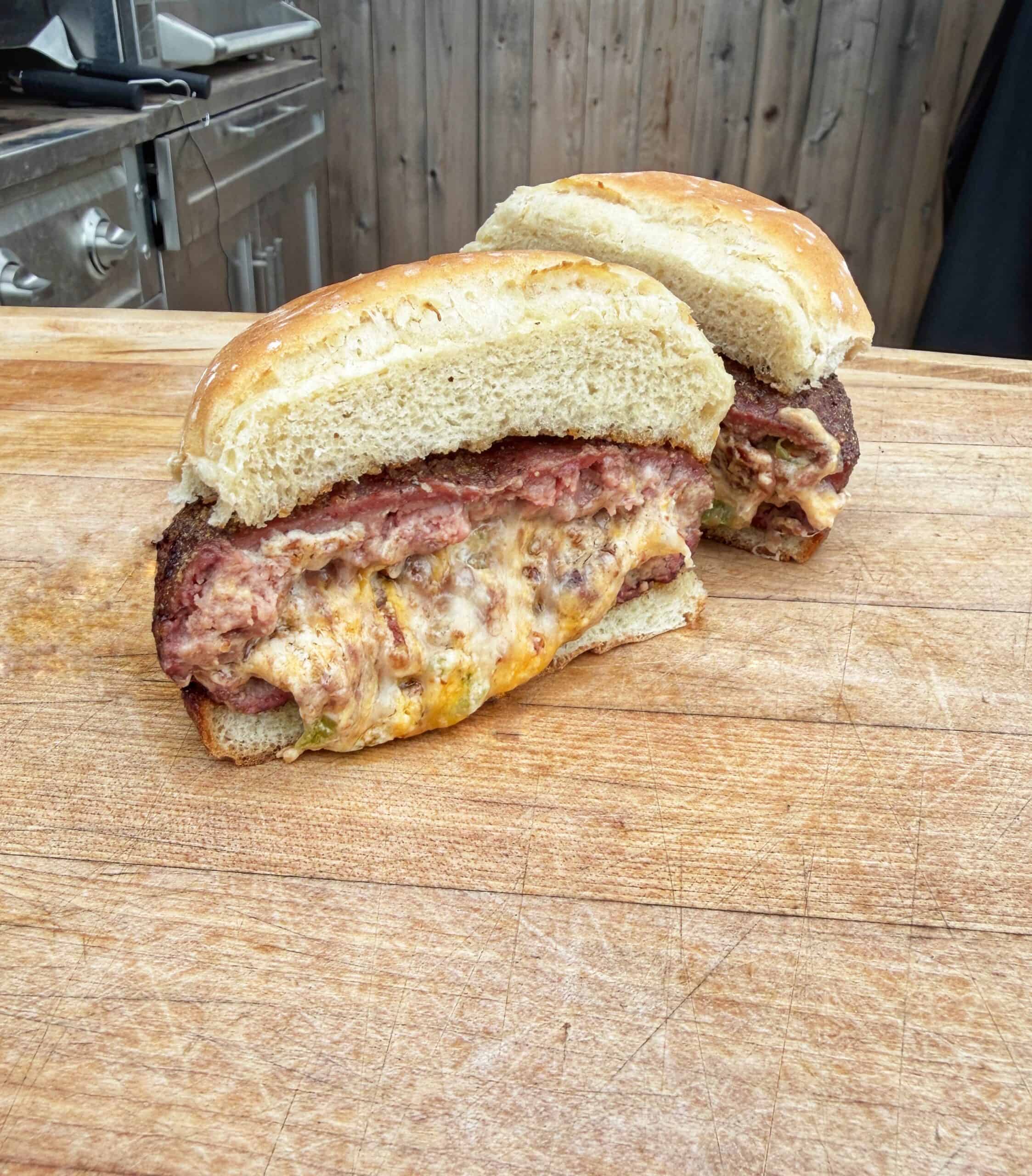 A large sandwich cut in half sits on a wooden cutting board, showing layers of melted cheese and meat inside a bun. A grill and wooden fence are visible in the background.