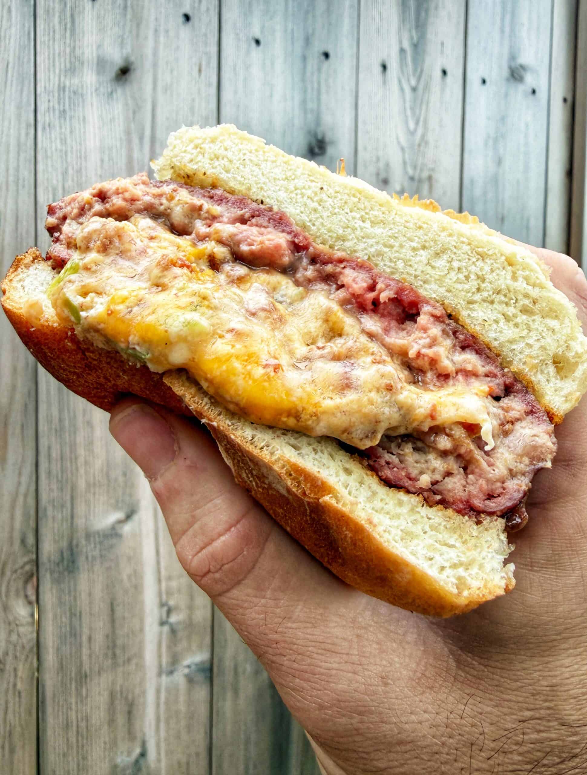 A hand holding a sandwich with a thick layer of ground meat and melted cheese inside a soft bun, with a wooden background.