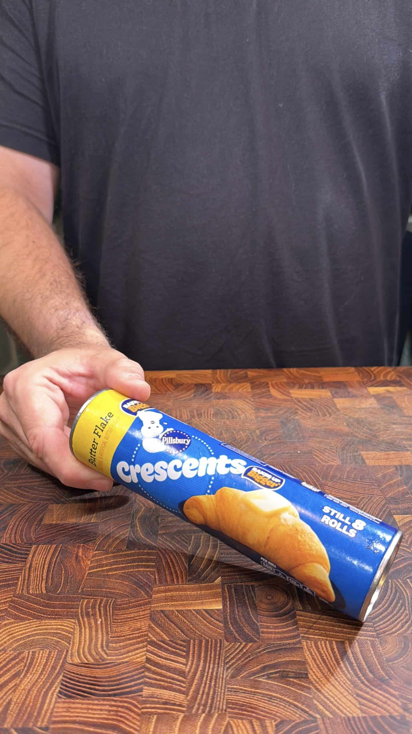 A person in a dark shirt holds a can of Pillsbury Crescents dough on a wooden patterned surface. The can is blue and yellow, showing an image of baked crescent rolls and the Pillsbury Doughboy.