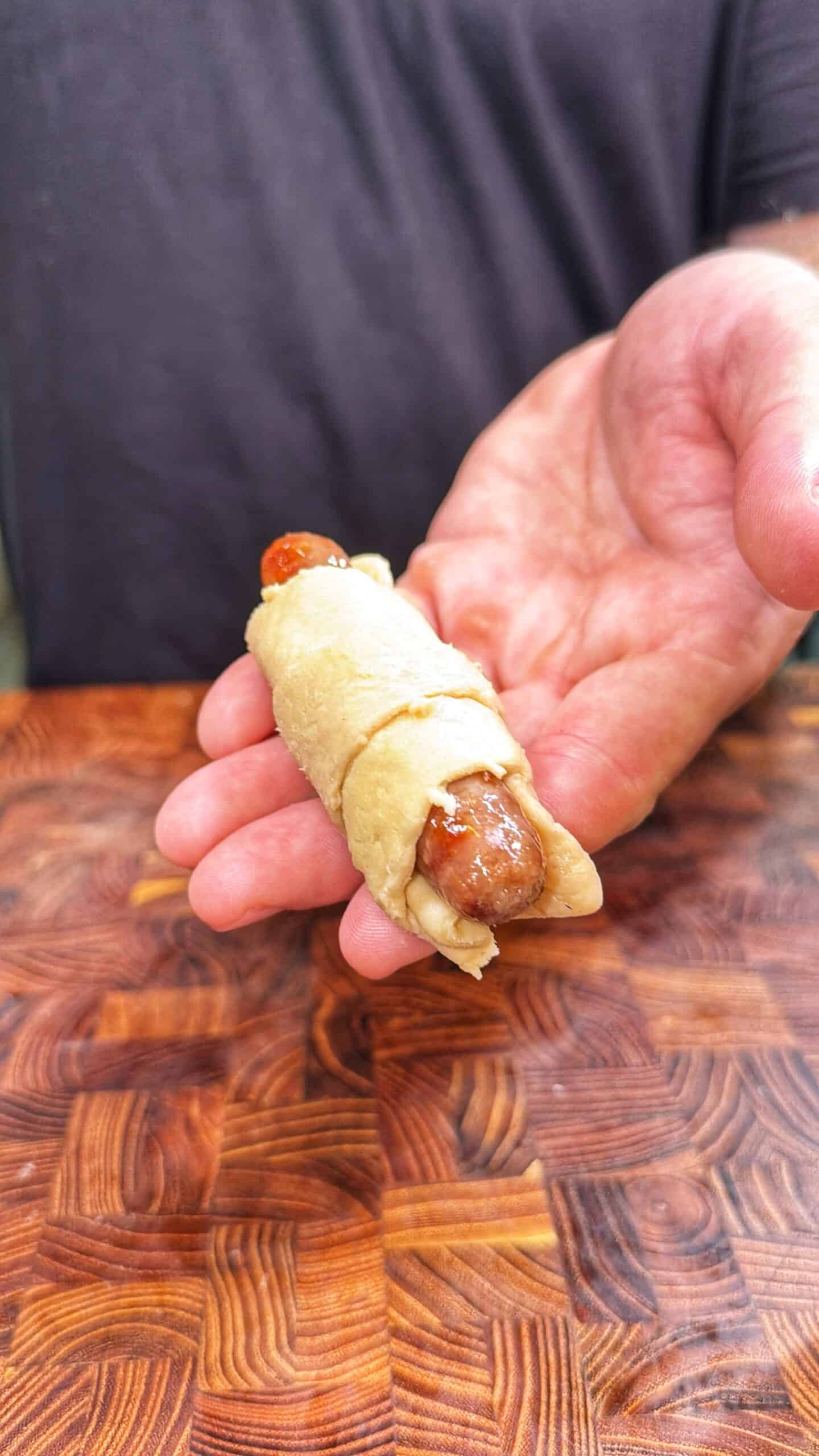 A hand holds a small sausage wrapped in dough, resembling a pig in a blanket, over a wooden surface.