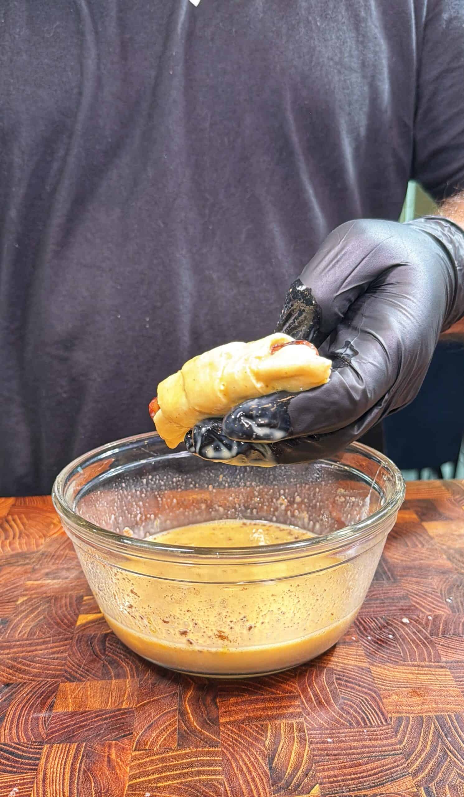 A person wearing a black glove holds a crab claw above a glass bowl filled with a light brown sauce on a wooden surface.