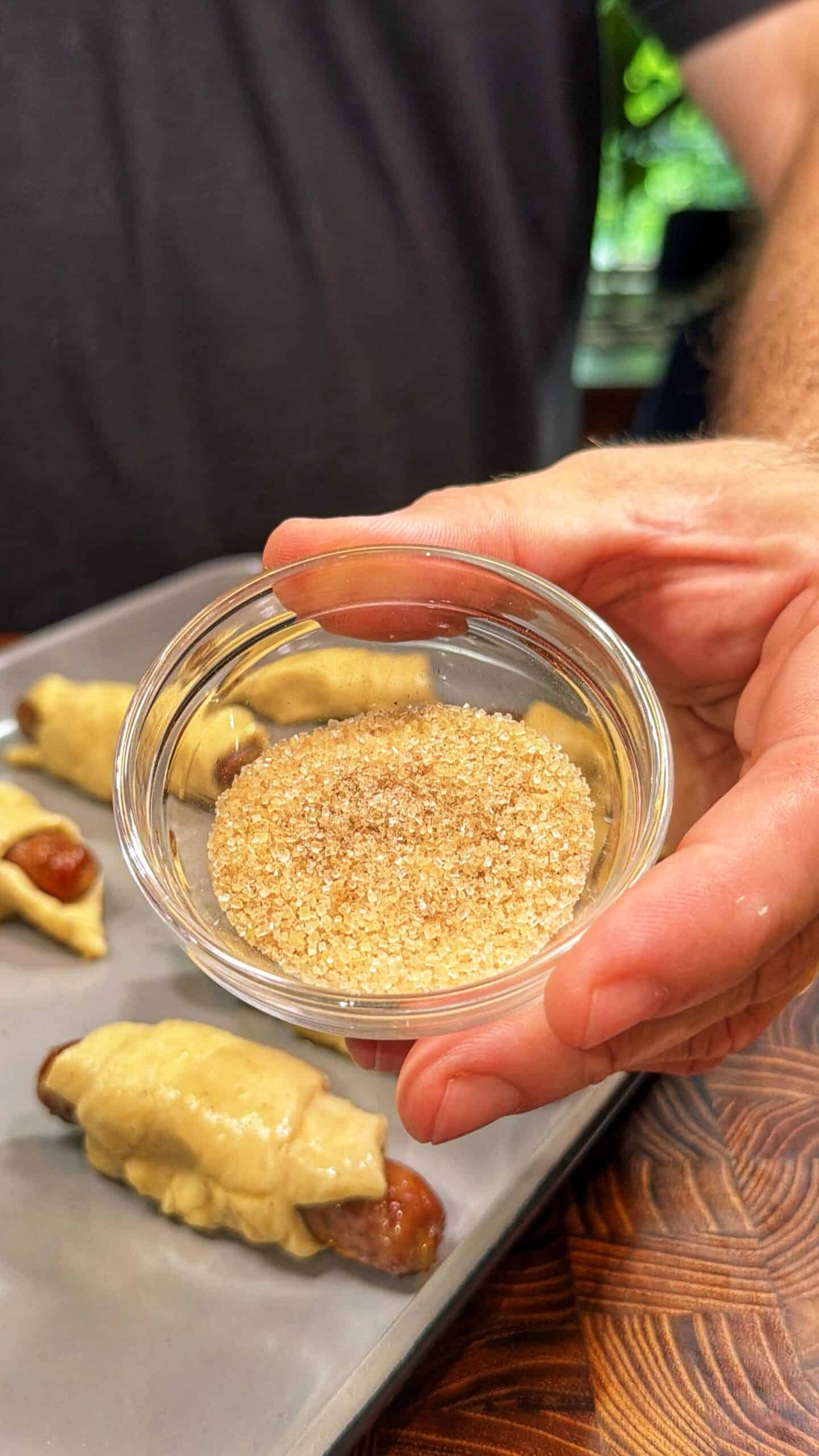 A hand holds a small glass bowl filled with coarse brown sugar over a tray of unbaked pastry-wrapped sausages on a wooden surface.