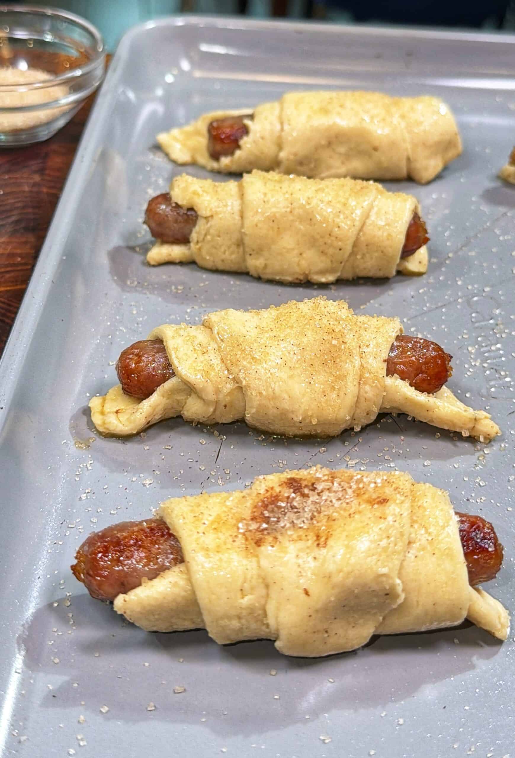 Three uncooked pigs in a blanket—sausages wrapped in dough—are placed on a baking sheet. The dough is sprinkled with seasoning, and a small bowl of seasoning is visible in the background.