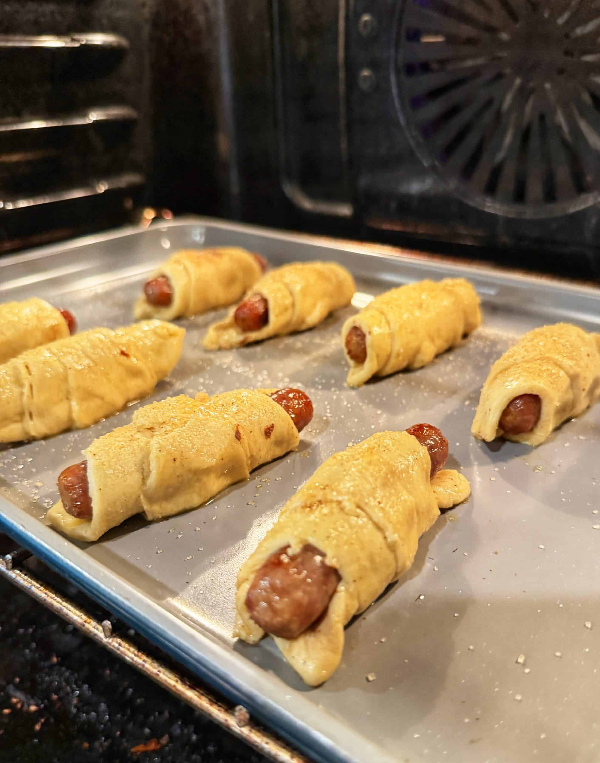 Unbaked pigs in a blanket wrapped in dough are arranged on a baking sheet inside an oven, ready to be baked.