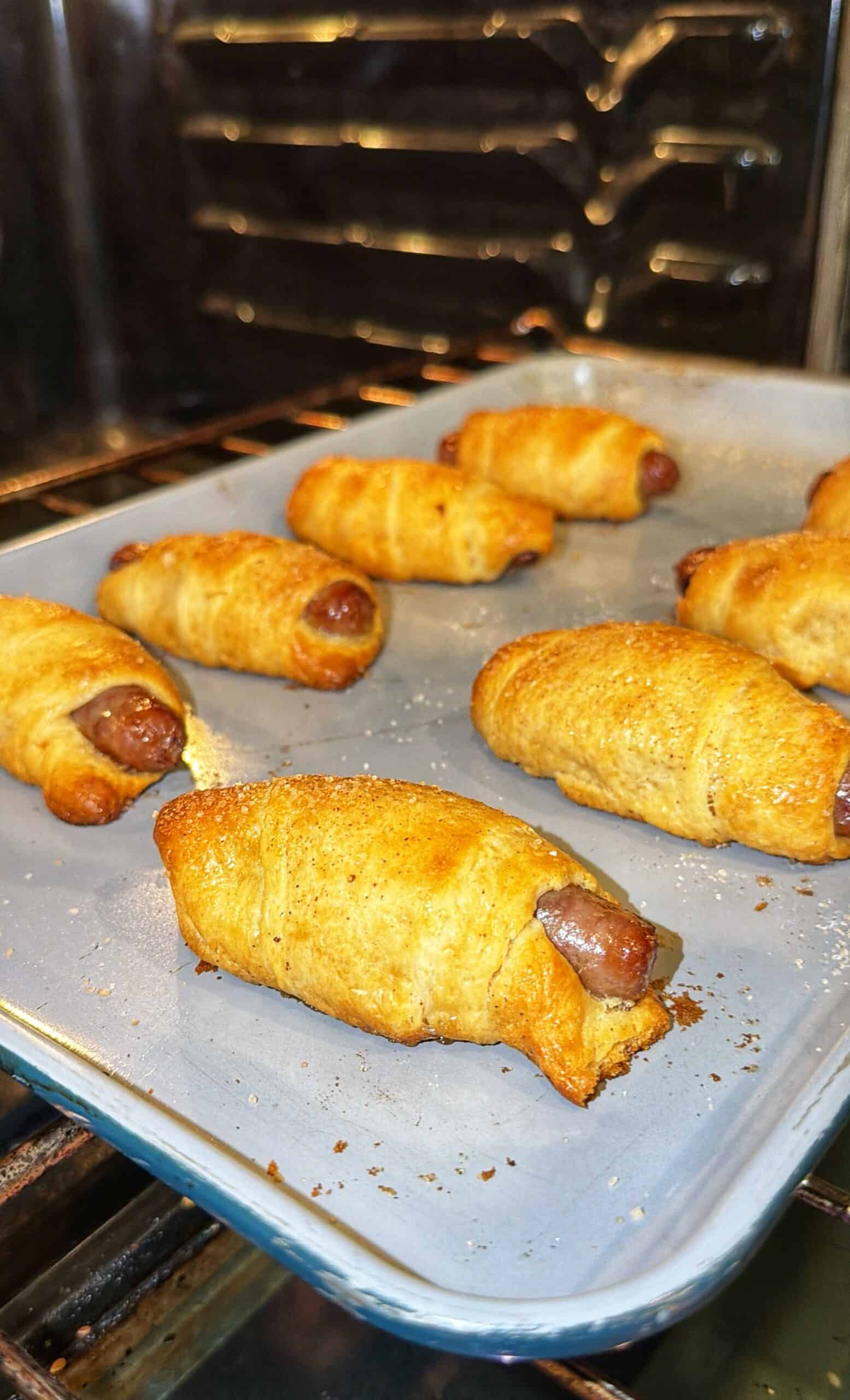 A baking tray with several golden-brown pigs in a blanket, small sausages wrapped in crescent dough, sits on an oven rack, with the oven interior visible in the background.