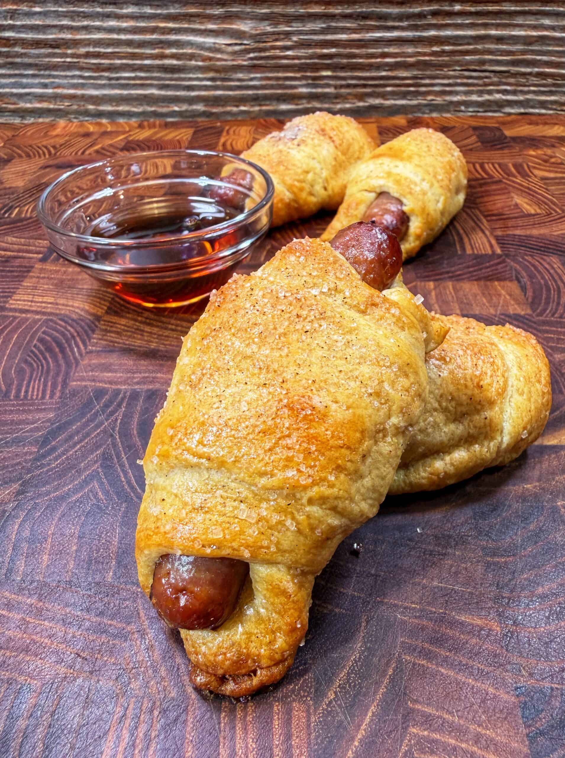 Golden brown sausage rolls wrapped in crescent dough are displayed on a wooden cutting board, with a small glass bowl of syrup for dipping in the background.