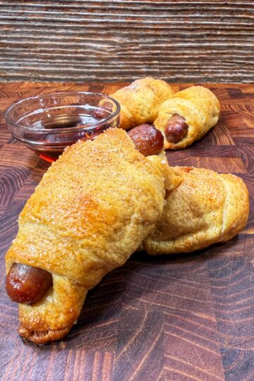 Three baked pigs in a blanket rest on a wooden surface, with a small glass bowl of dipping sauce in the background. The pastries are golden brown and slightly sugared.