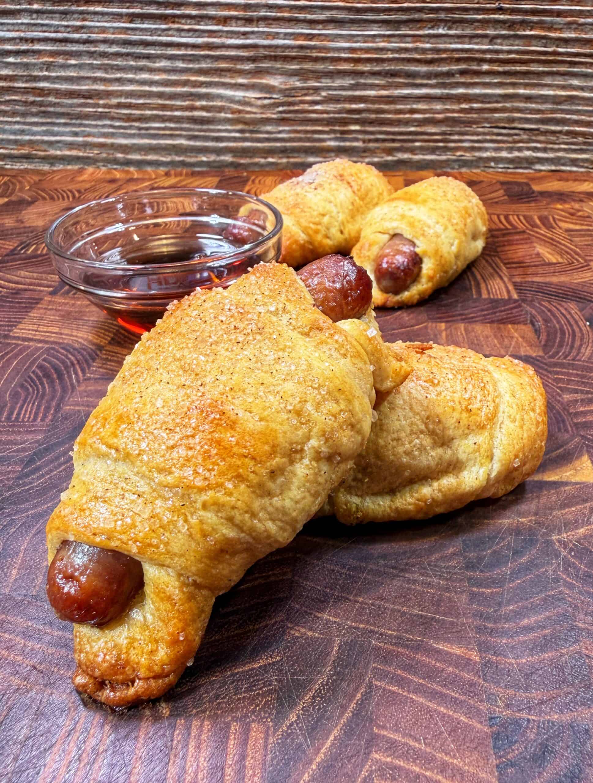 Three baked pigs in a blanket rest on a wooden surface, with a small glass bowl of dipping sauce in the background. The pastries are golden brown and slightly sugared.