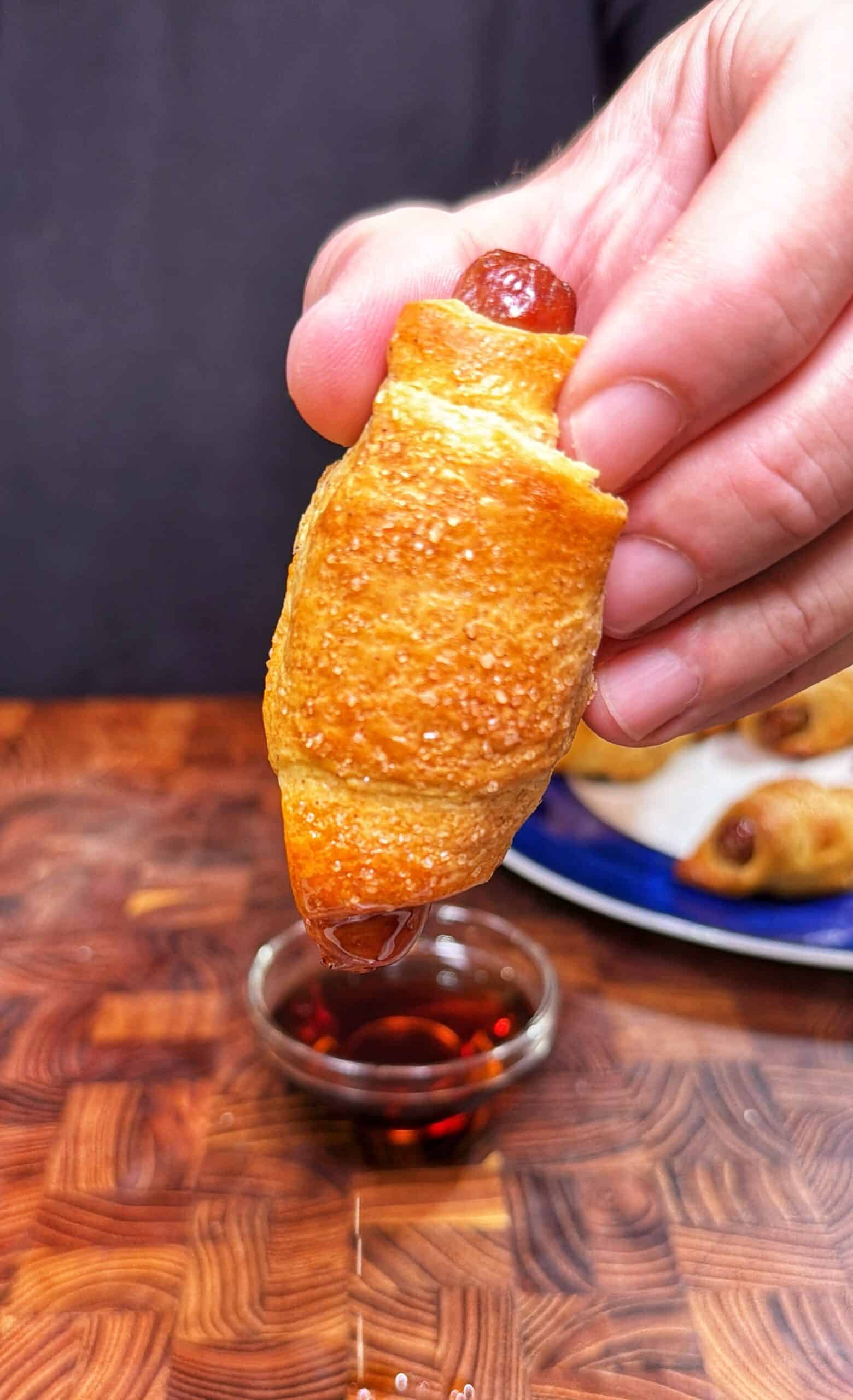 A hand holds a mini crescent roll-wrapped sausage, dipping it into a small glass bowl of sauce. A plate with more crescent roll sausages is seen in the background on a wooden surface.