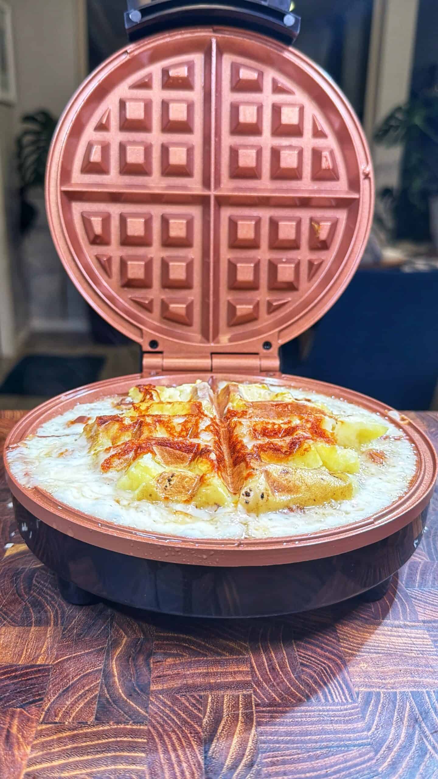 A round waffle maker with its lid open reveals a golden, cheesy dish divided into quarters, with melted cheese bubbling over the edges, sitting on a wooden surface.