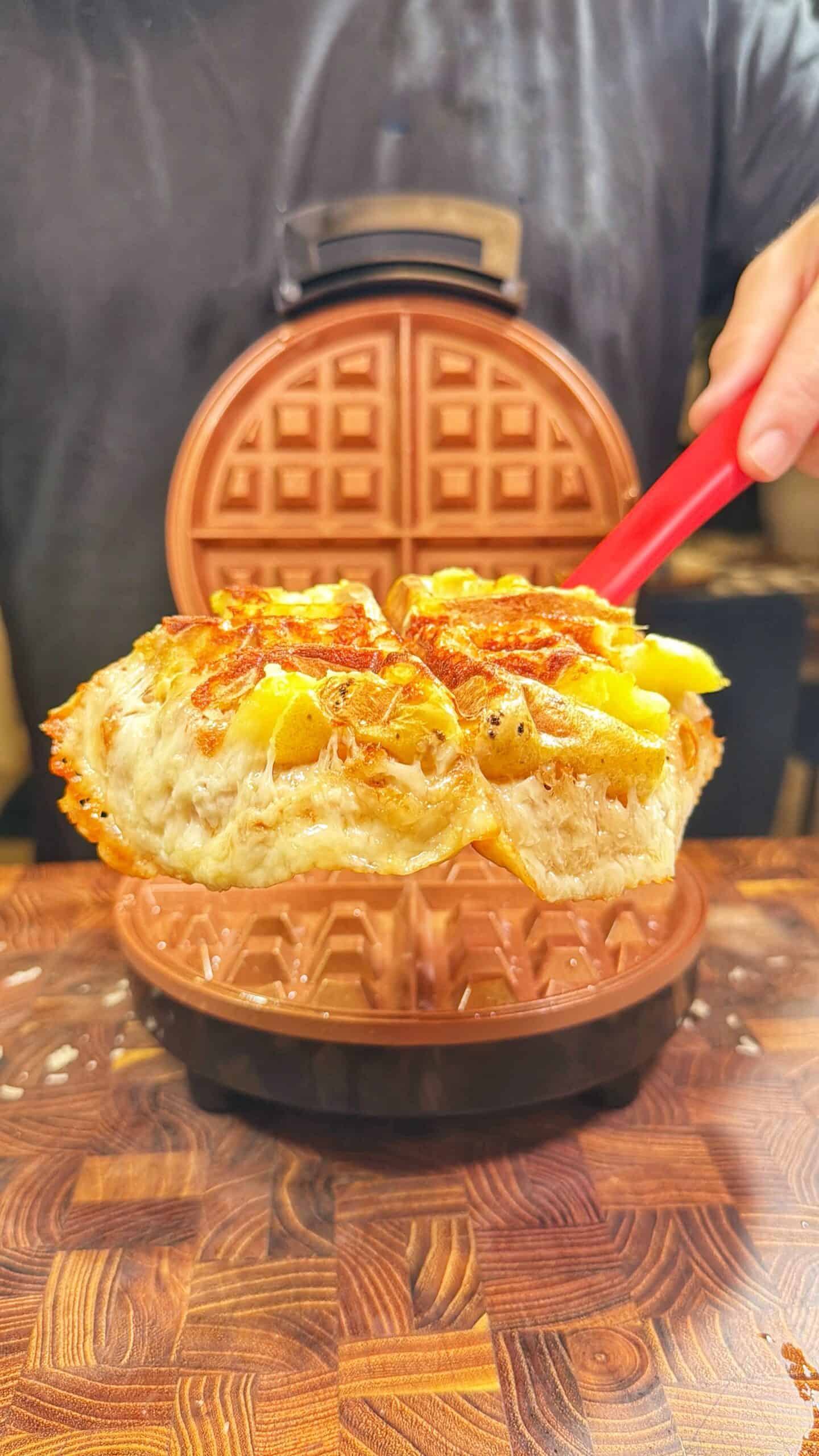 A person lifts a cooked, cheesy waffle with pieces of potato from an open waffle maker using a red spatula. The waffle maker sits on a wooden surface.