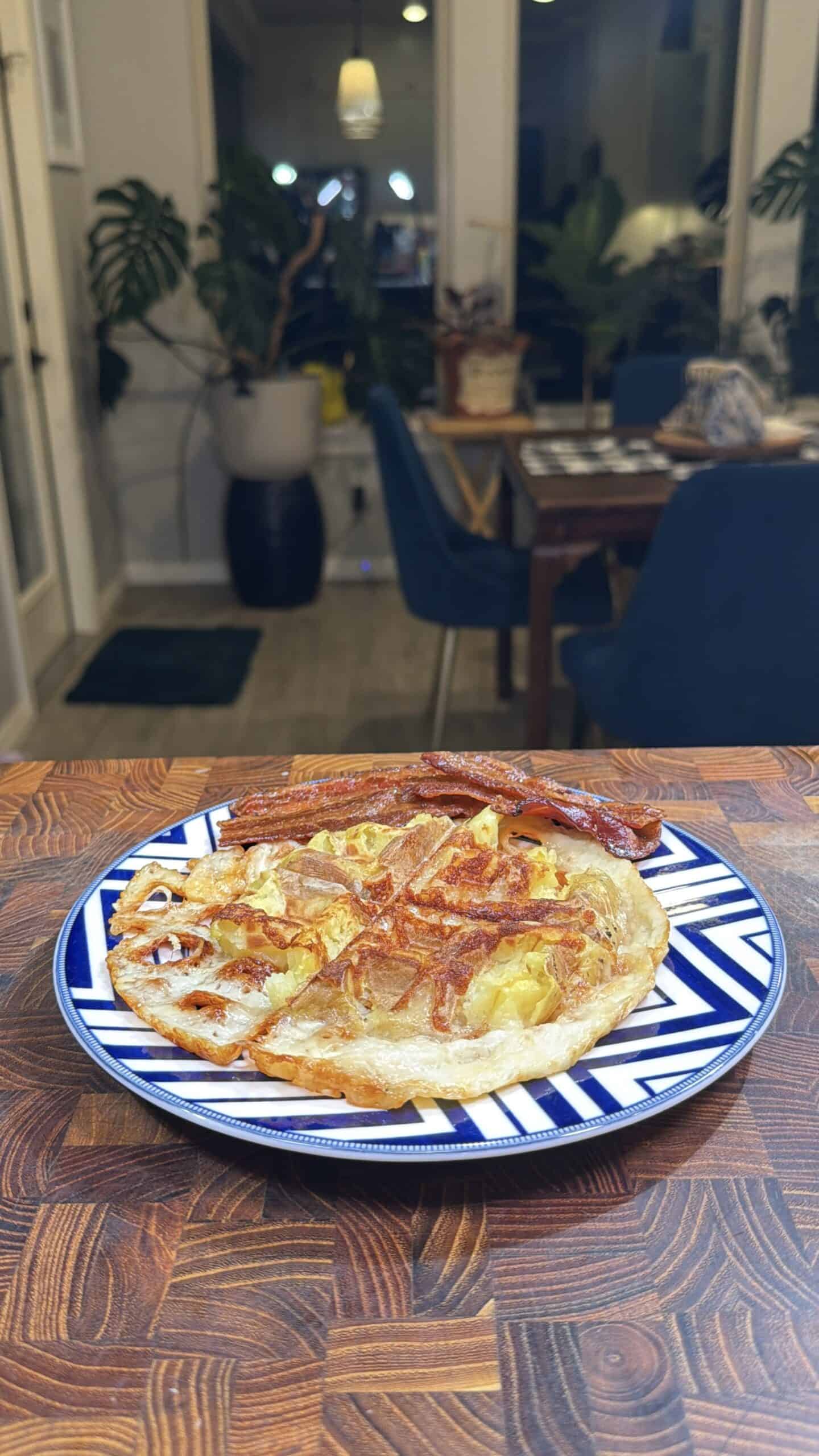 A plate with a homemade pizza topped with cheese and bacon sits on a wooden table. In the background, a dining area with blue chairs, plants, and a table is visible.