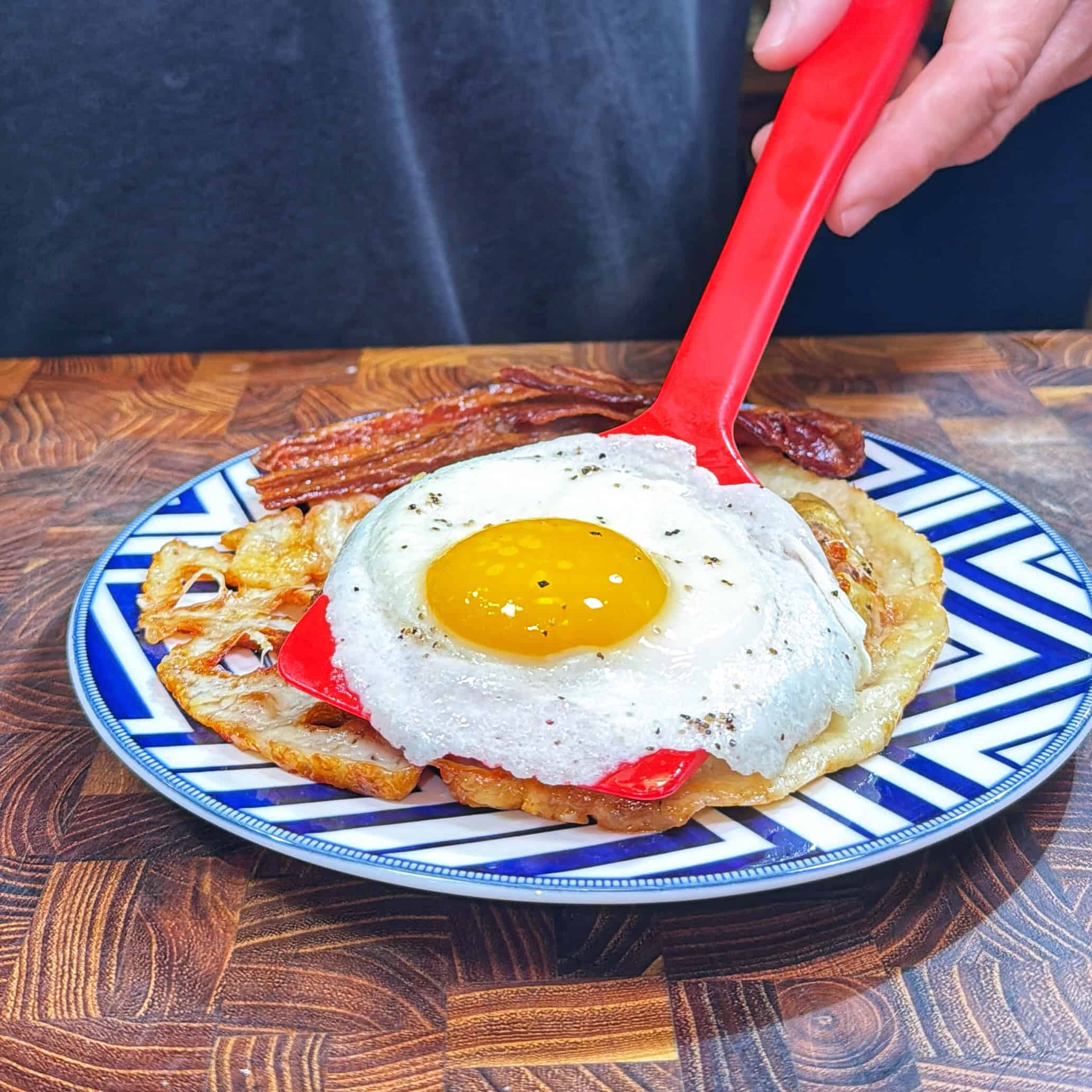 A person uses a red spatula to serve a sunny-side-up egg on top of a crispy lotus root slice, with bacon strips on a blue and white patterned plate on a wooden surface.