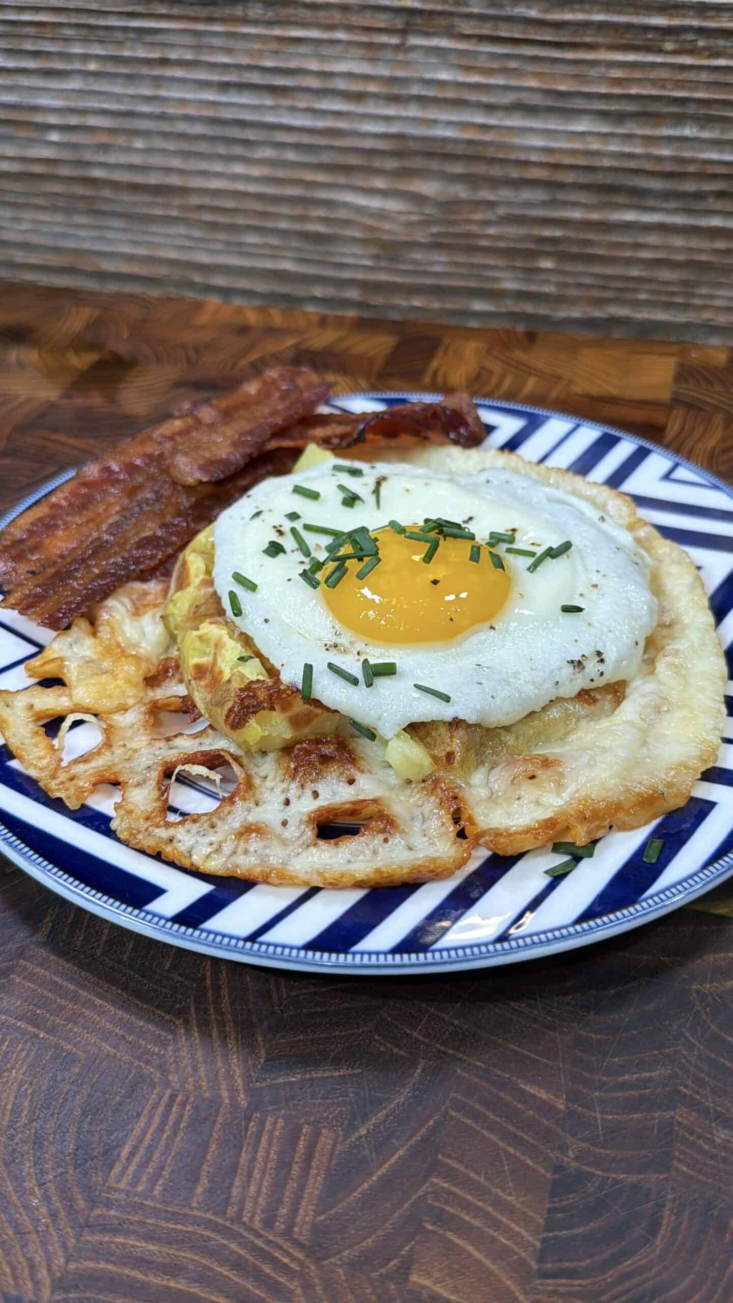 A striped plate holds a crispy fried egg topped with chopped chives, resting on a slice of melted cheese and fried lotus root, with two strips of cooked bacon on the side.