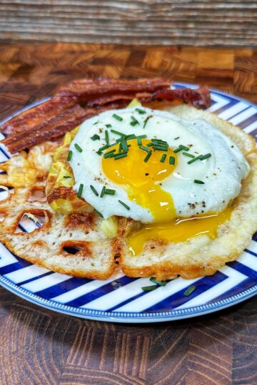 A plate with a sunny side up egg topped with chopped chives, crispy bacon strips, and fried lotus root, served on a blue and white patterned dish on a wooden surface.