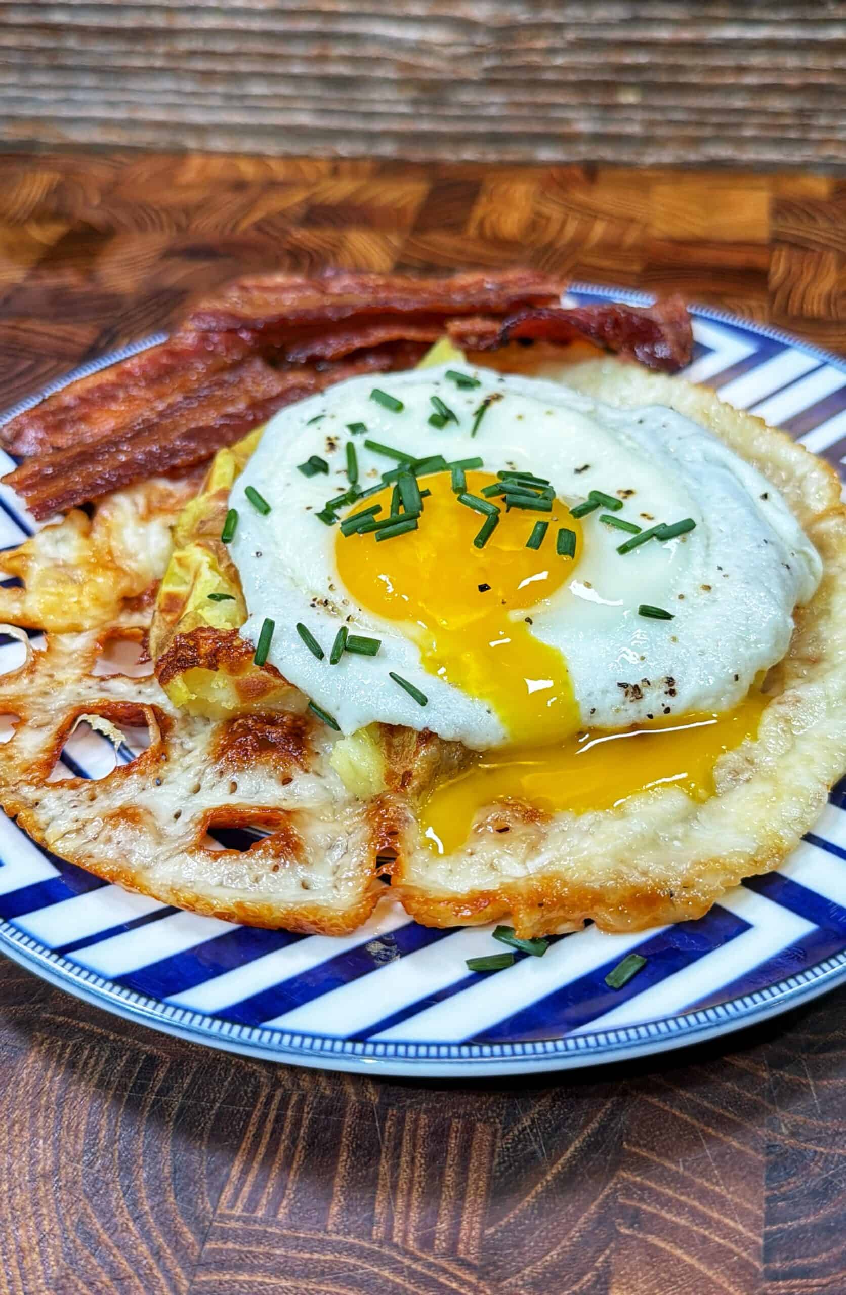 A plate with a sunny side up egg topped with chopped chives, crispy bacon strips, and fried lotus root, served on a blue and white patterned dish on a wooden surface.