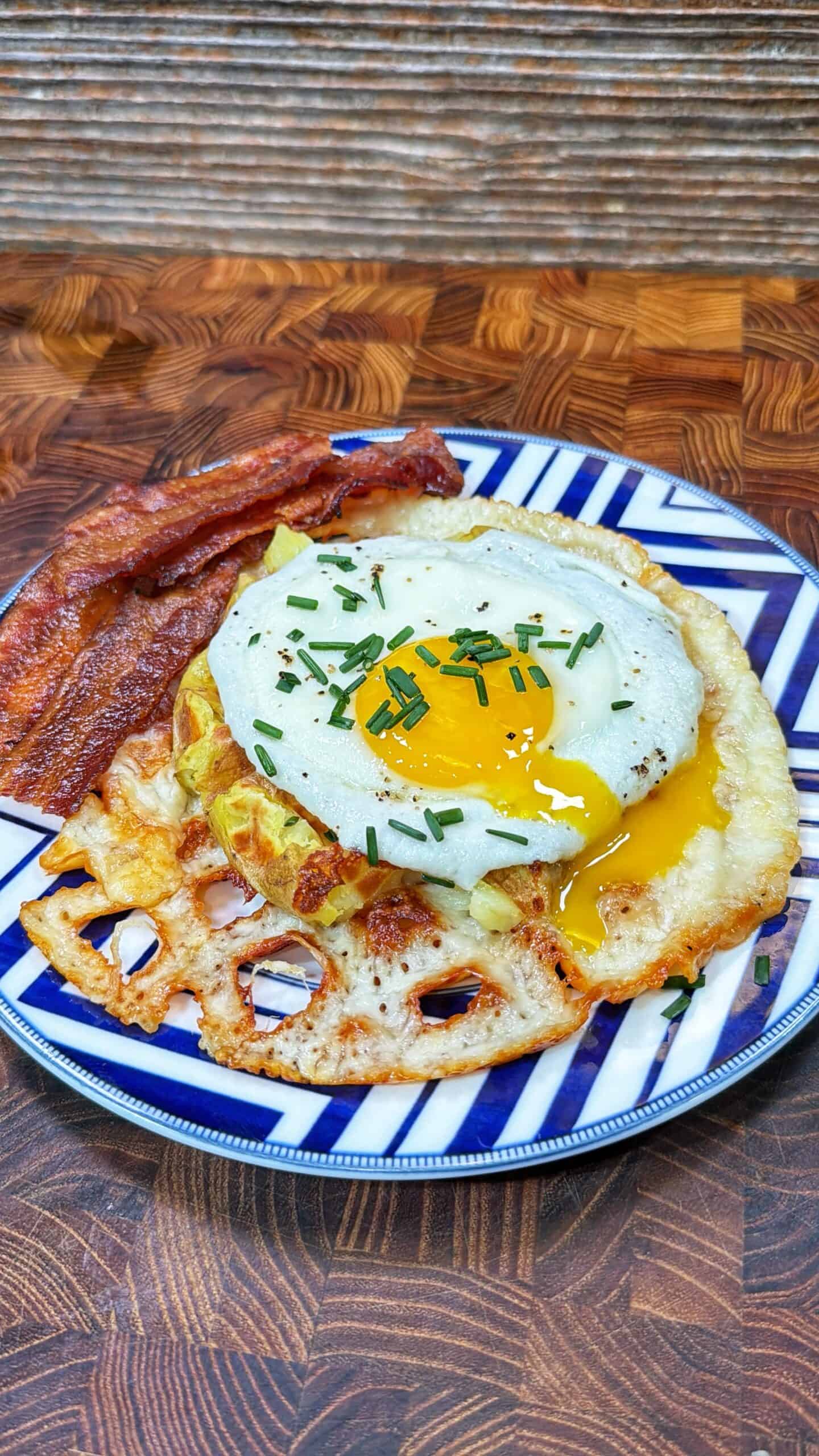 A blue and white plate with crispy bacon strips, a sunny-side-up egg topped with chopped chives, and golden brown potato waffles on a wooden surface.