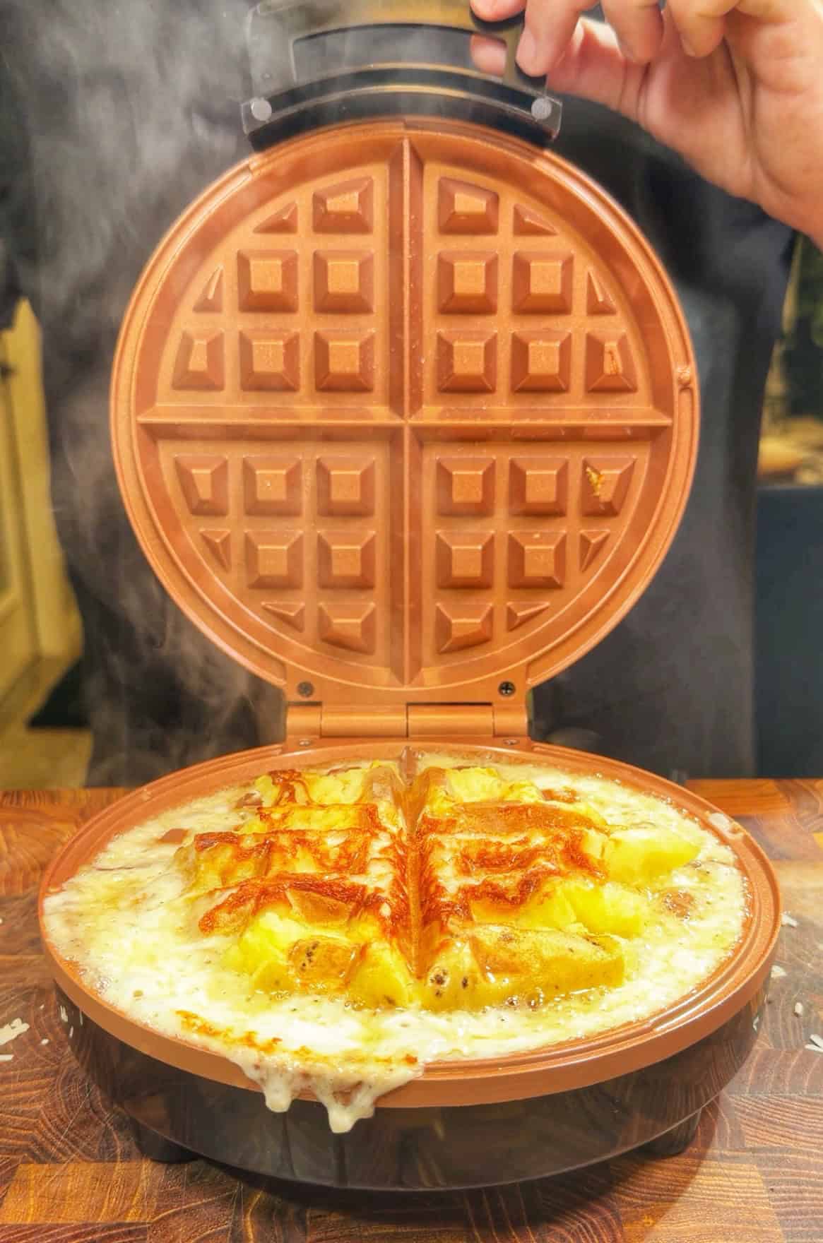 A golden, cheesy waffle melts and bubbles in a waffle maker, with steam rising and cheese oozing over the sides. A hand is lifting the lid to reveal the hot, freshly cooked waffle.