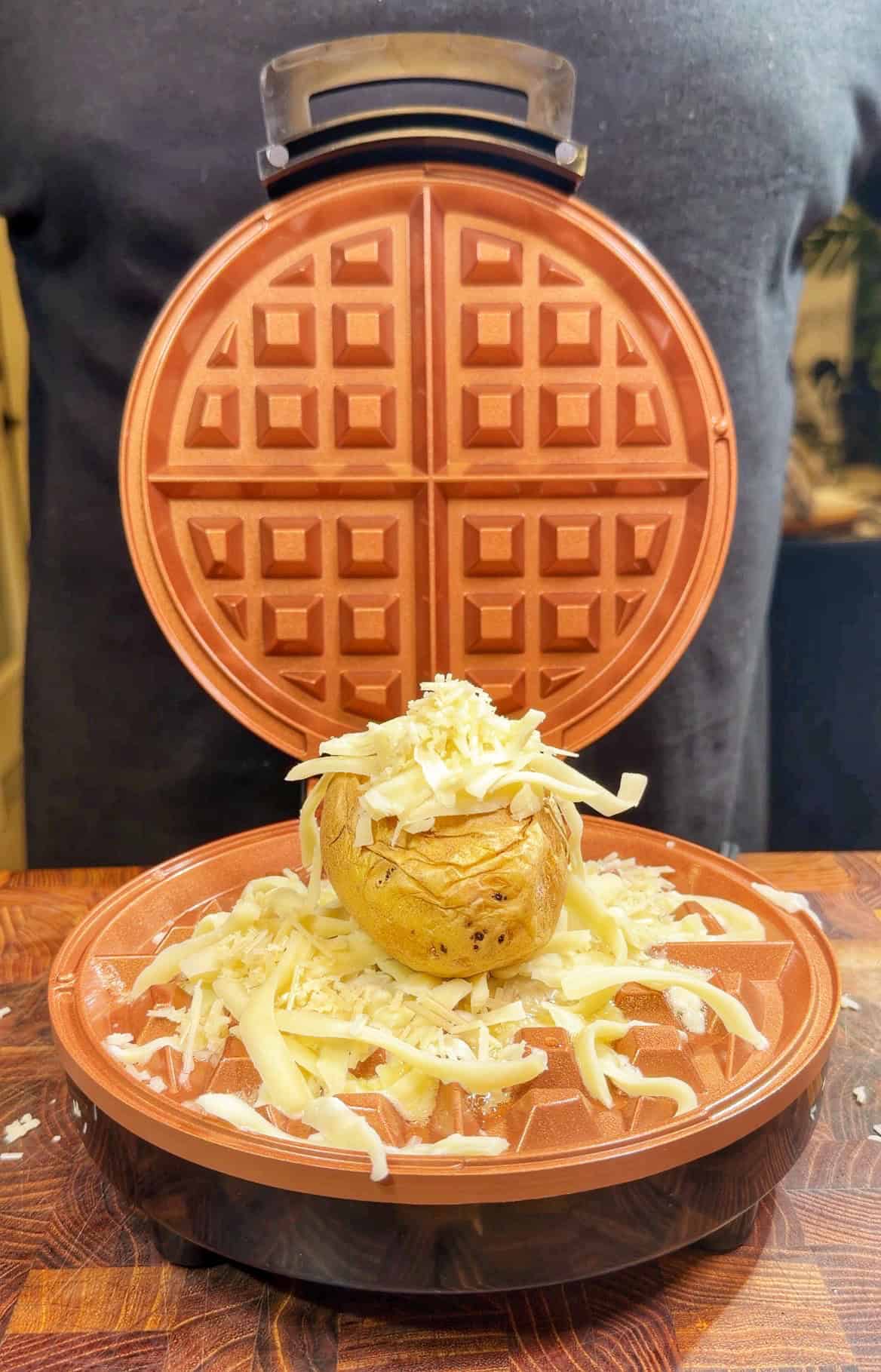 A whole baked potato topped with shredded cheese sits on the cooking plate of an open waffle maker, ready to be pressed, with a person standing in the background.