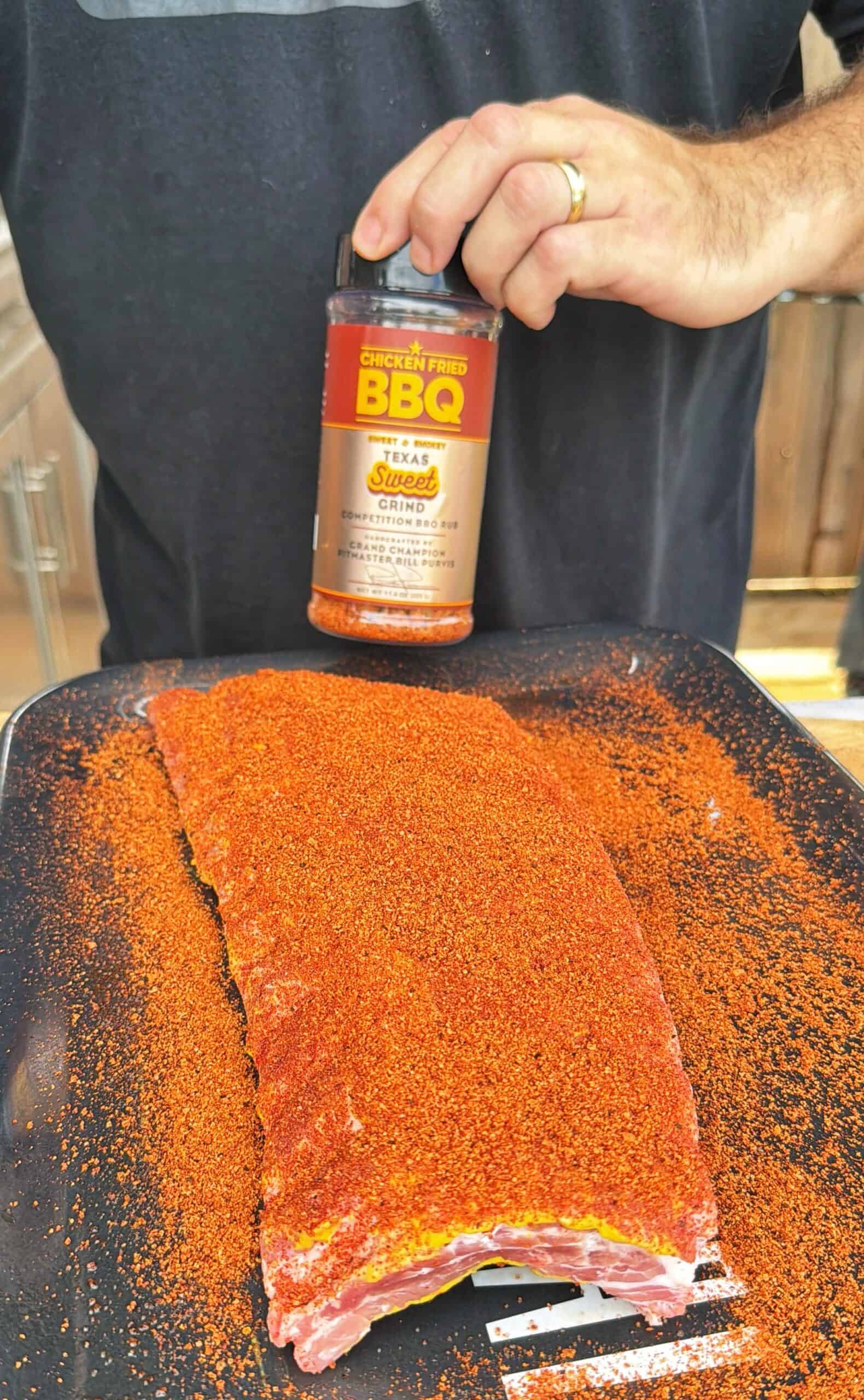 A person sprinkles BBQ rub seasoning from a container onto a rack of raw ribs covered in a reddish spice rub, placed on a black tray. The person is wearing a dark shirt and a wedding ring.