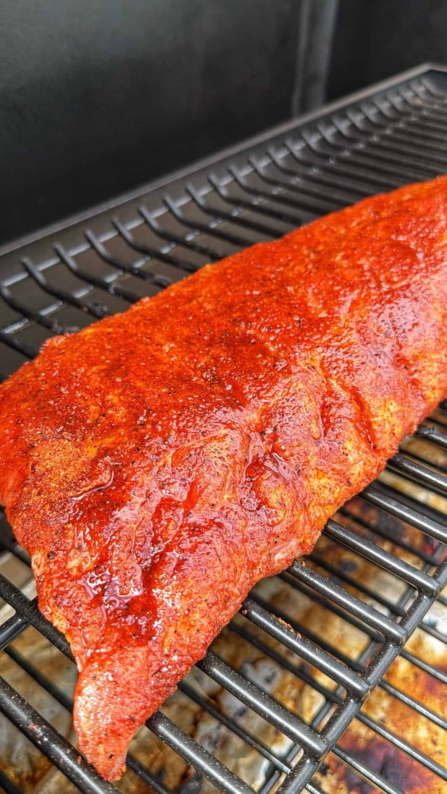 A close-up of a rack of ribs coated in a reddish seasoning rub, cooking on a grill grate inside a barbecue smoker.