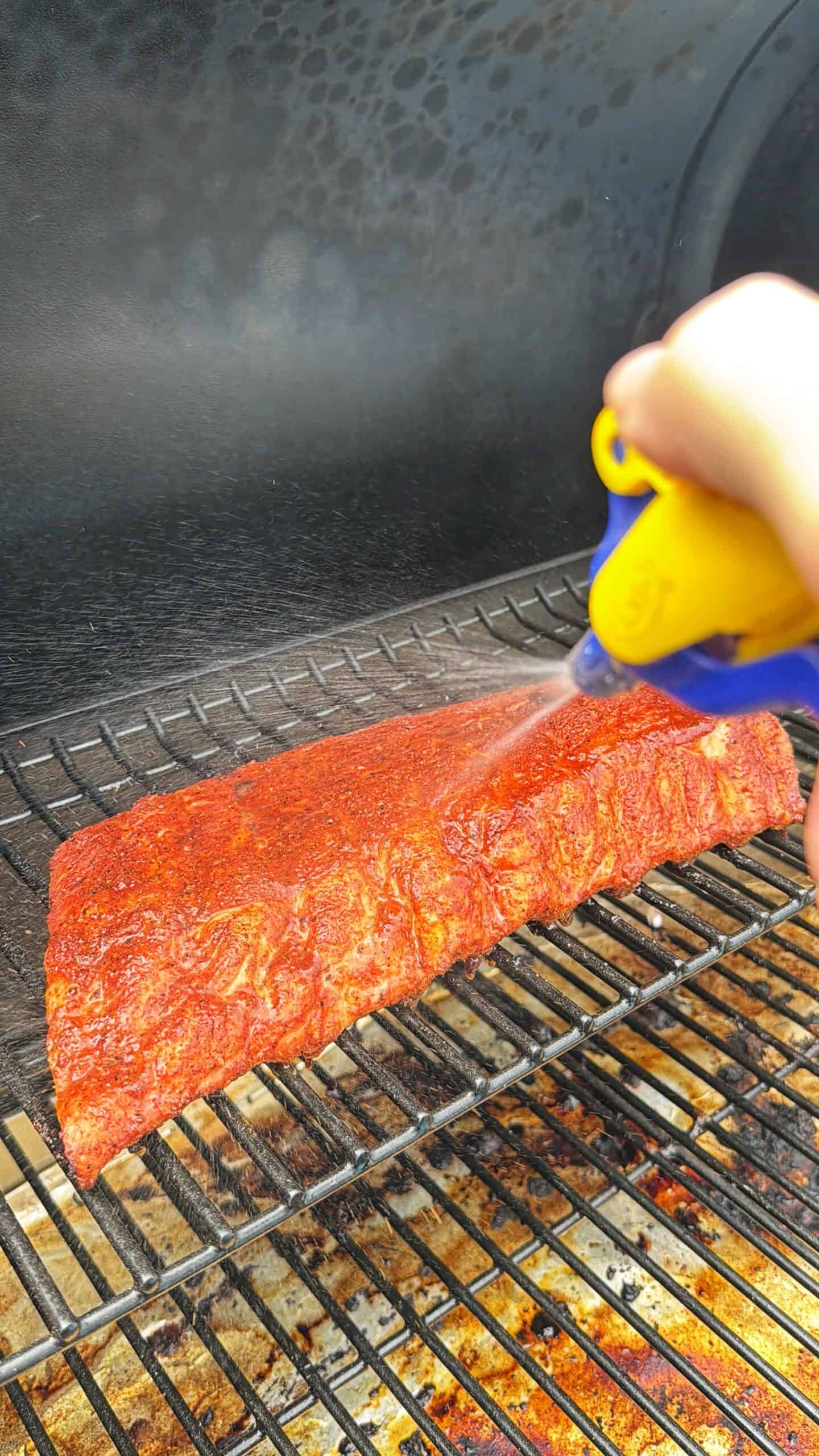A hand uses a spray bottle to mist a rack of seasoned ribs cooking on a grill, helping keep the meat moist during the smoking or grilling process.