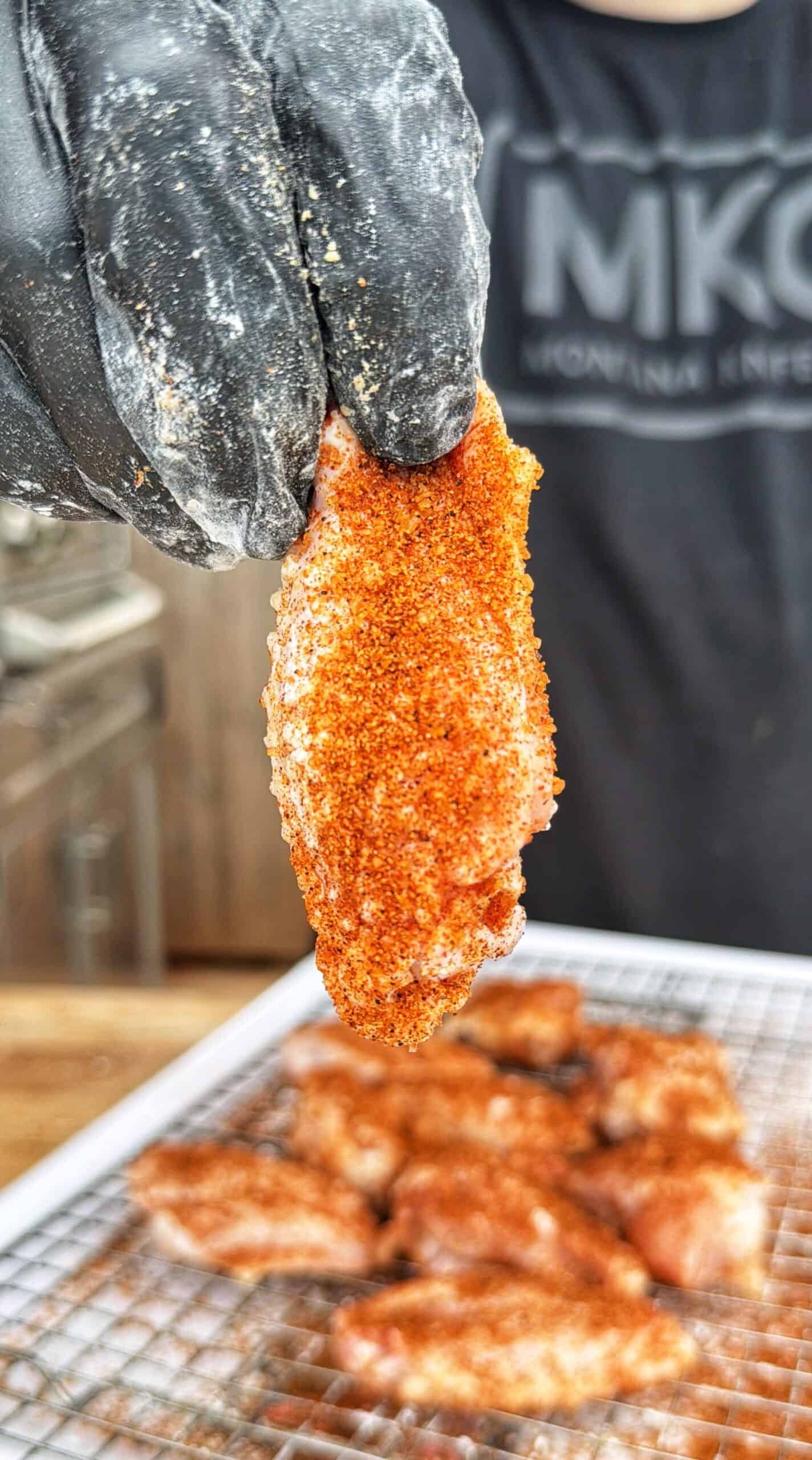 A gloved hand holds up a seasoned chicken wing over a wire rack with more seasoned wings below, ready for cooking.