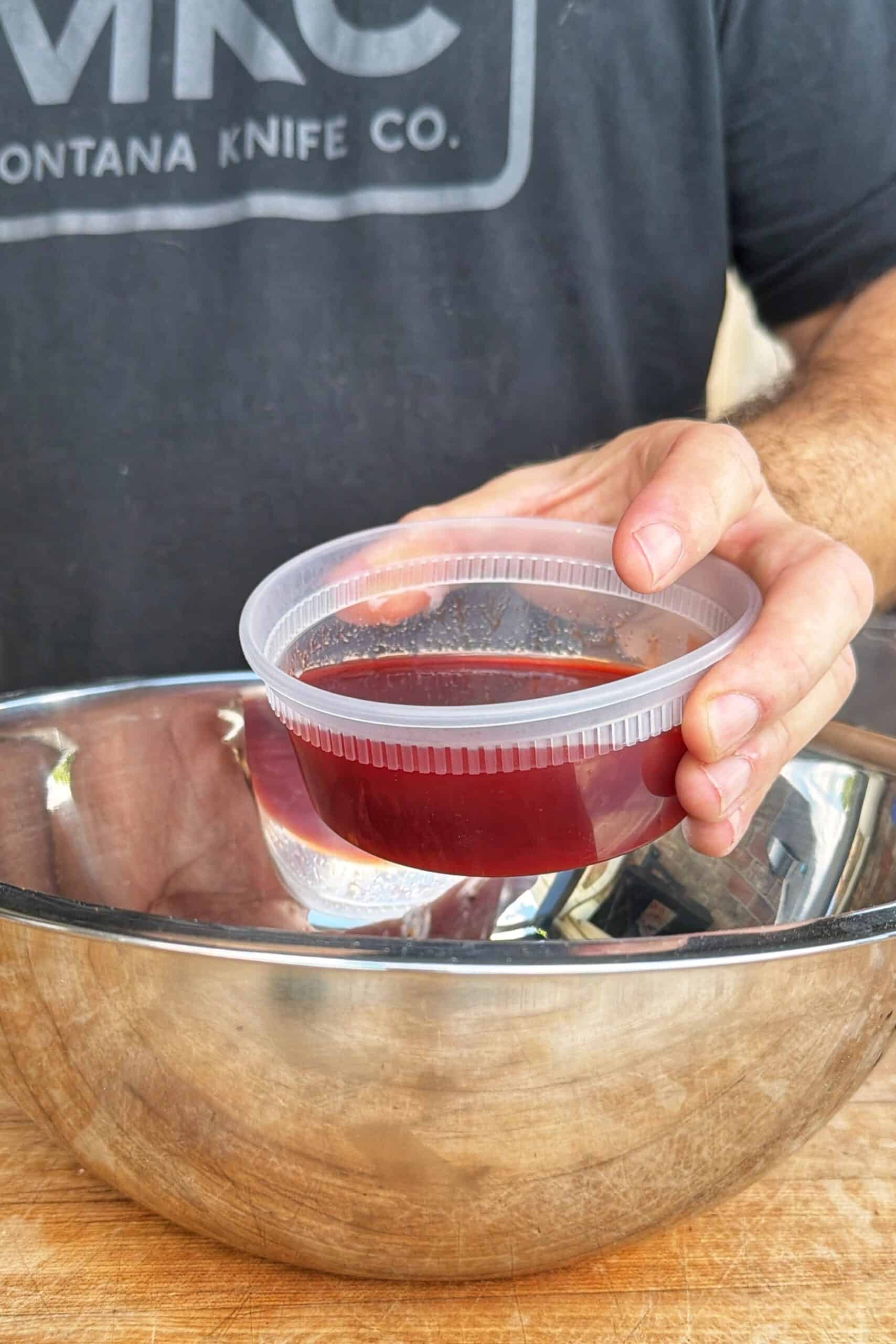 A person holds a small plastic container filled with a dark red liquid above a large metal mixing bowl on a wooden surface. The person is wearing a dark shirt with Montana Knife Co. partially visible.