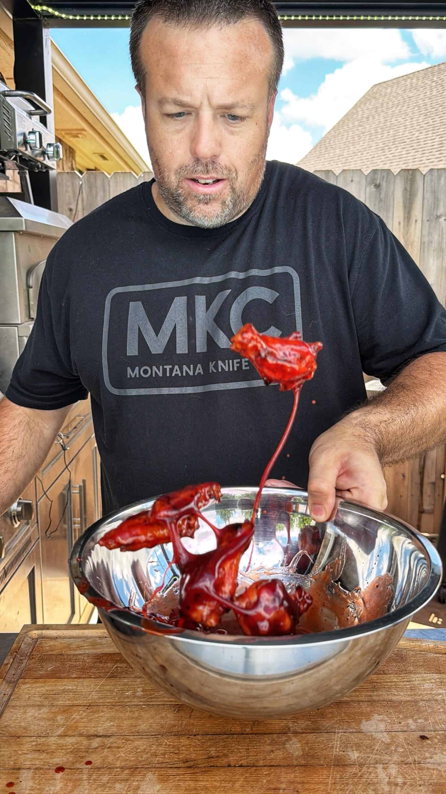 A man in a black Montana Knife t-shirt stands outdoors, holding a metal bowl filled with bright red sauced chicken wings, one of which is being lifted out, dripping sauce.