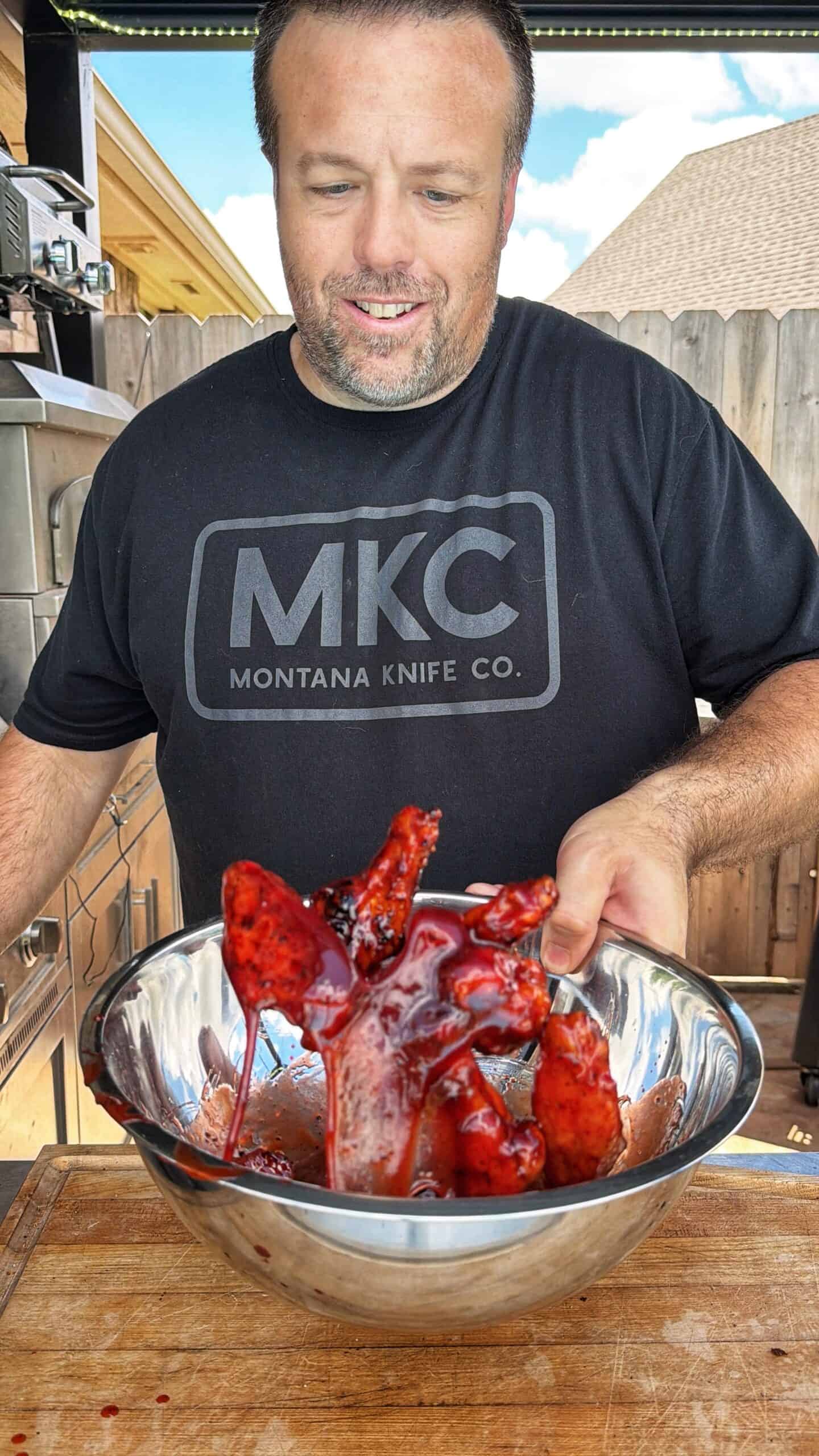 A man wearing a black Montana Knife Co. shirt holds a metal bowl filled with raw, marinated meat coated in a red sauce, standing outdoors near a wooden fence.