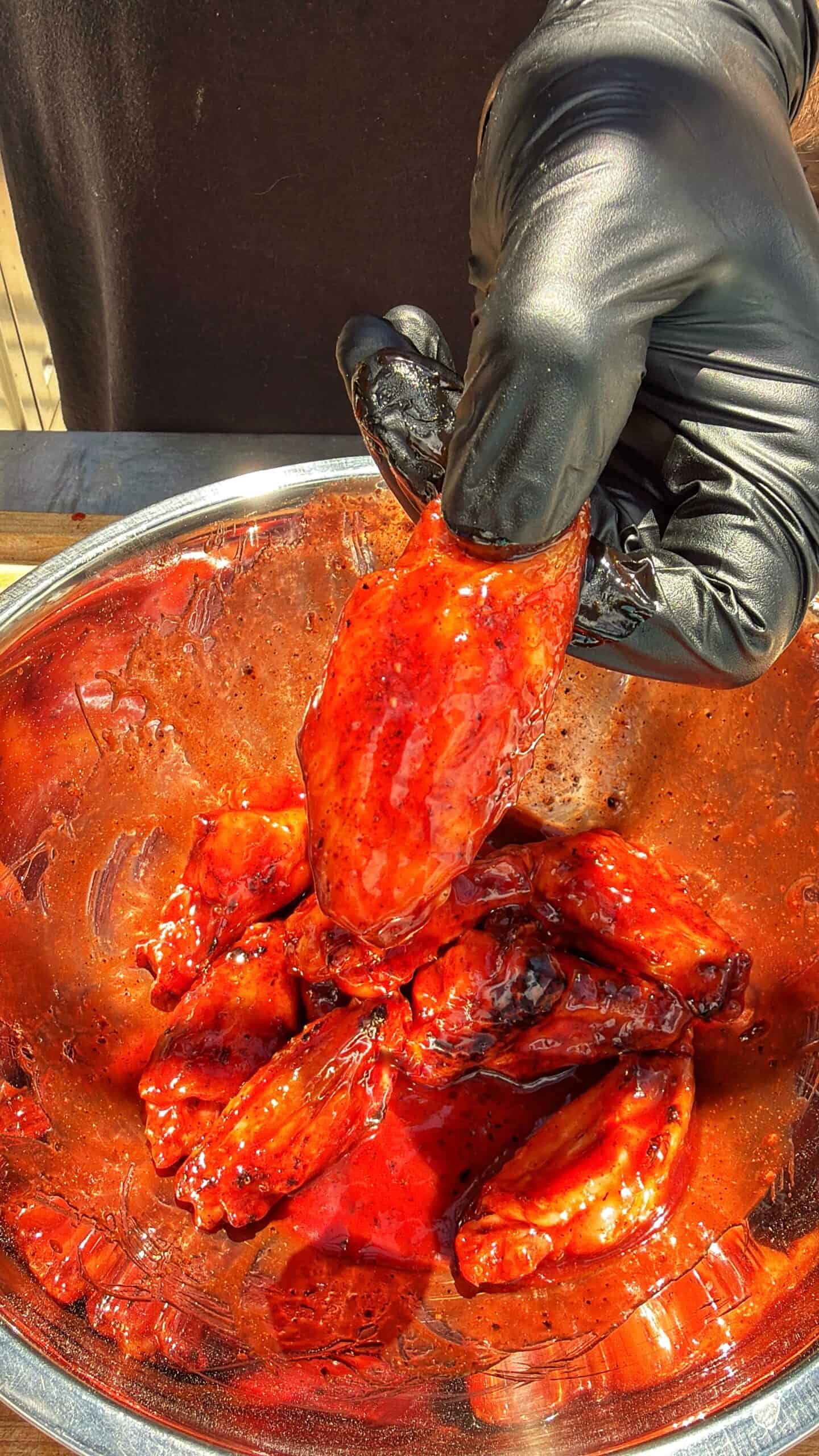 A hand wearing a black glove holds a saucy, red chicken wing above a metal bowl filled with more glazed wings. The wings are coated in a shiny, thick sauce, and the background is out of focus.