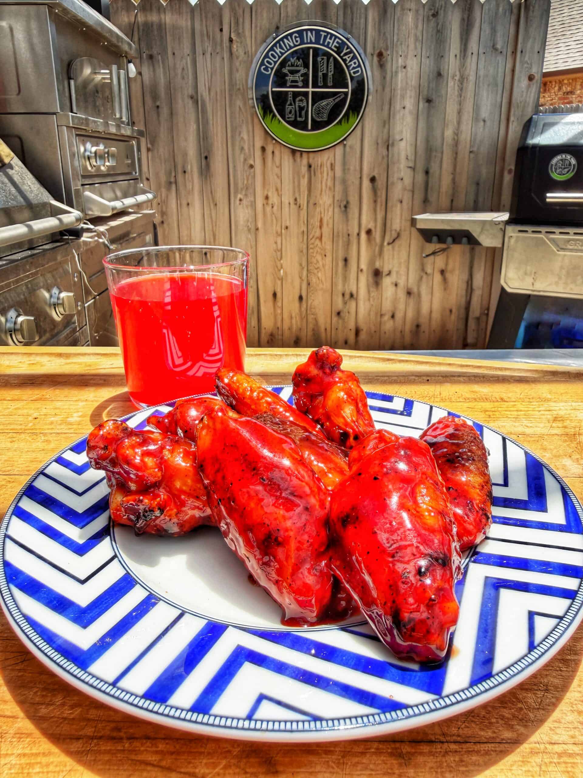 A plate of bright red grilled chicken wings sits on a blue and white patterned dish next to a glass of red drink, set on a wooden counter in an outdoor kitchen with grills and a fence in the background.