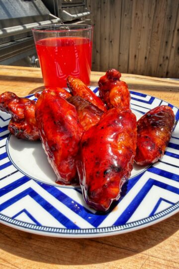 A blue and white plate with six glazed barbecue chicken wings sits on a wooden table. Behind the plate is a glass of bright red drink. A grill and wooden fence are visible in the background.