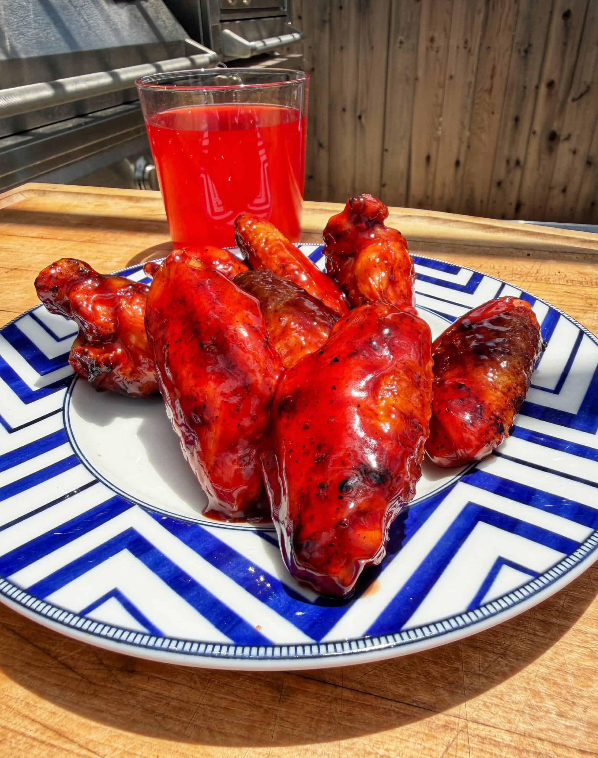 A blue and white plate with six glazed barbecue chicken wings sits on a wooden table. Behind the plate is a glass of bright red drink. A grill and wooden fence are visible in the background.
