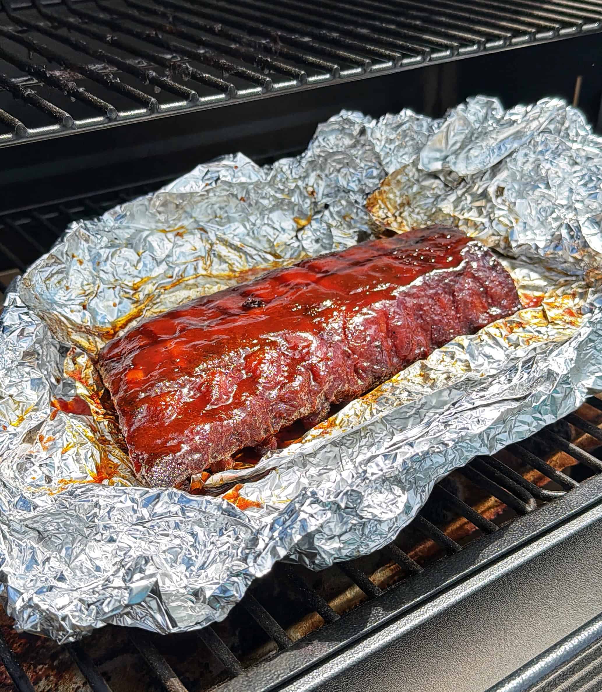 A rack of glazed barbecue ribs sits on crumpled aluminum foil inside a grill, with shiny sauce coating the meat and grill grates visible above and below.