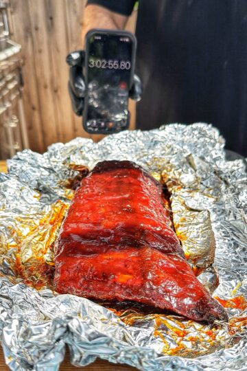 A slab of glazed barbecue ribs rests on crumpled aluminum foil, while a person in black holds up a phone displaying a timer reading 3:02:55.80 in the background.