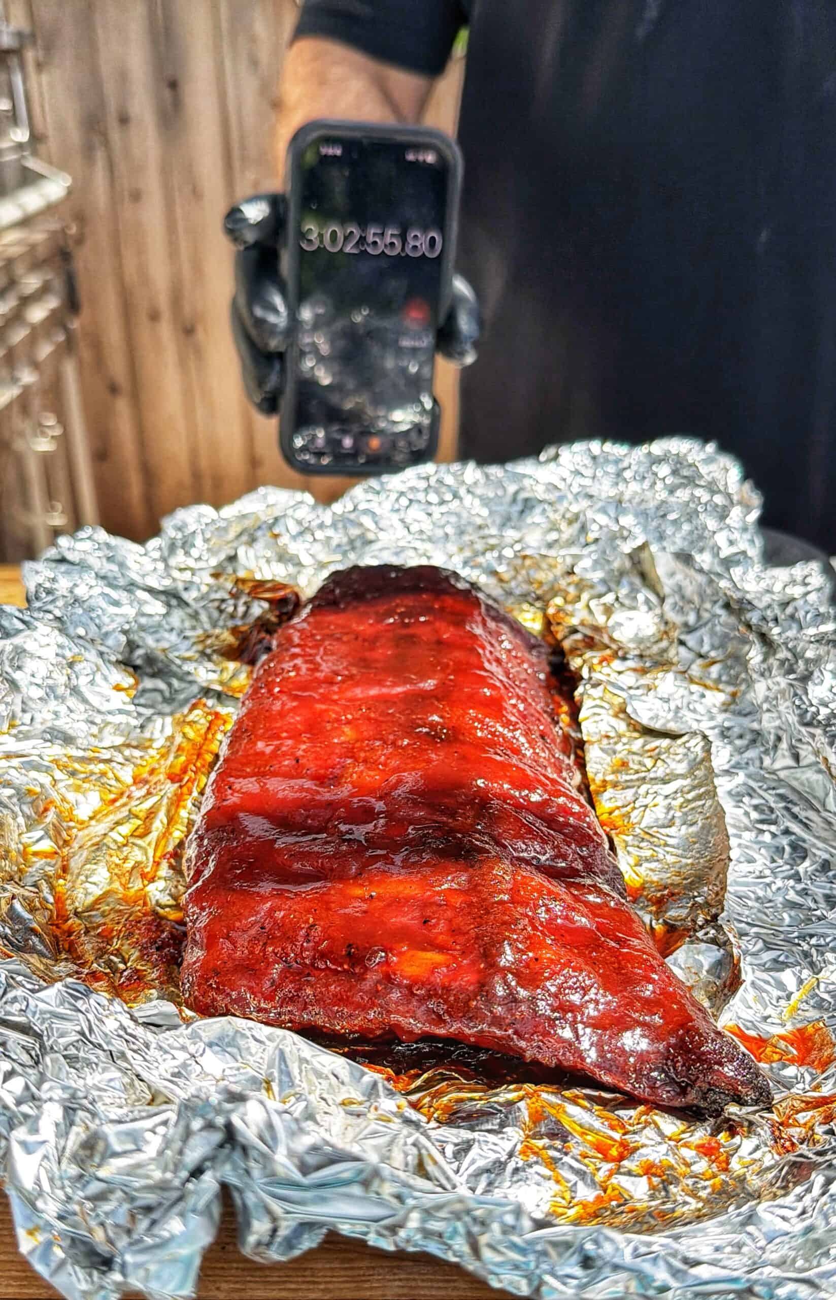 A slab of glazed barbecue ribs rests on crumpled aluminum foil, while a person in black holds up a phone displaying a timer reading 3:02:55.80 in the background.