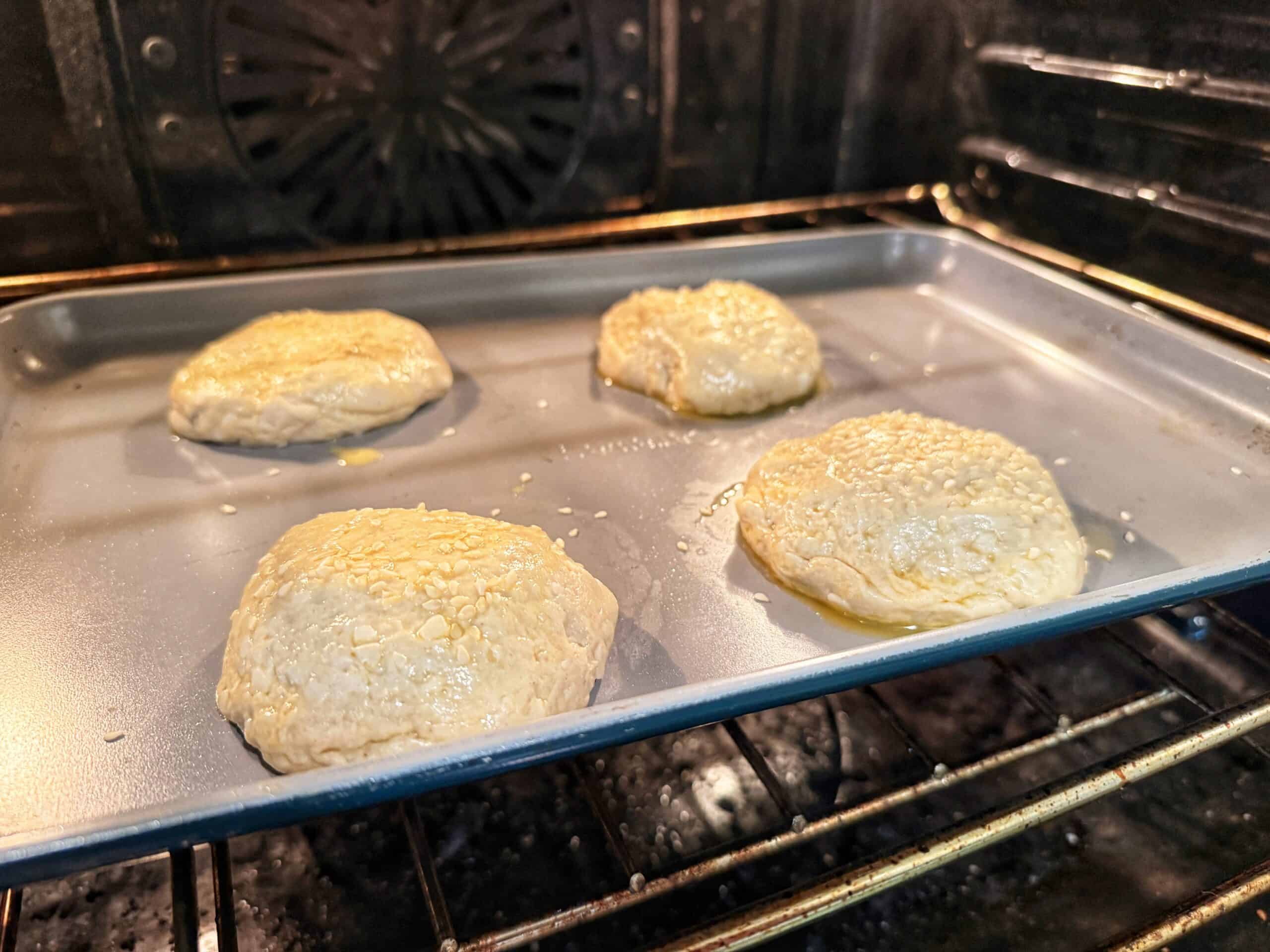Four unbaked, round dough buns topped with sesame seeds are on a baking sheet inside an oven, beginning to rise and bake.