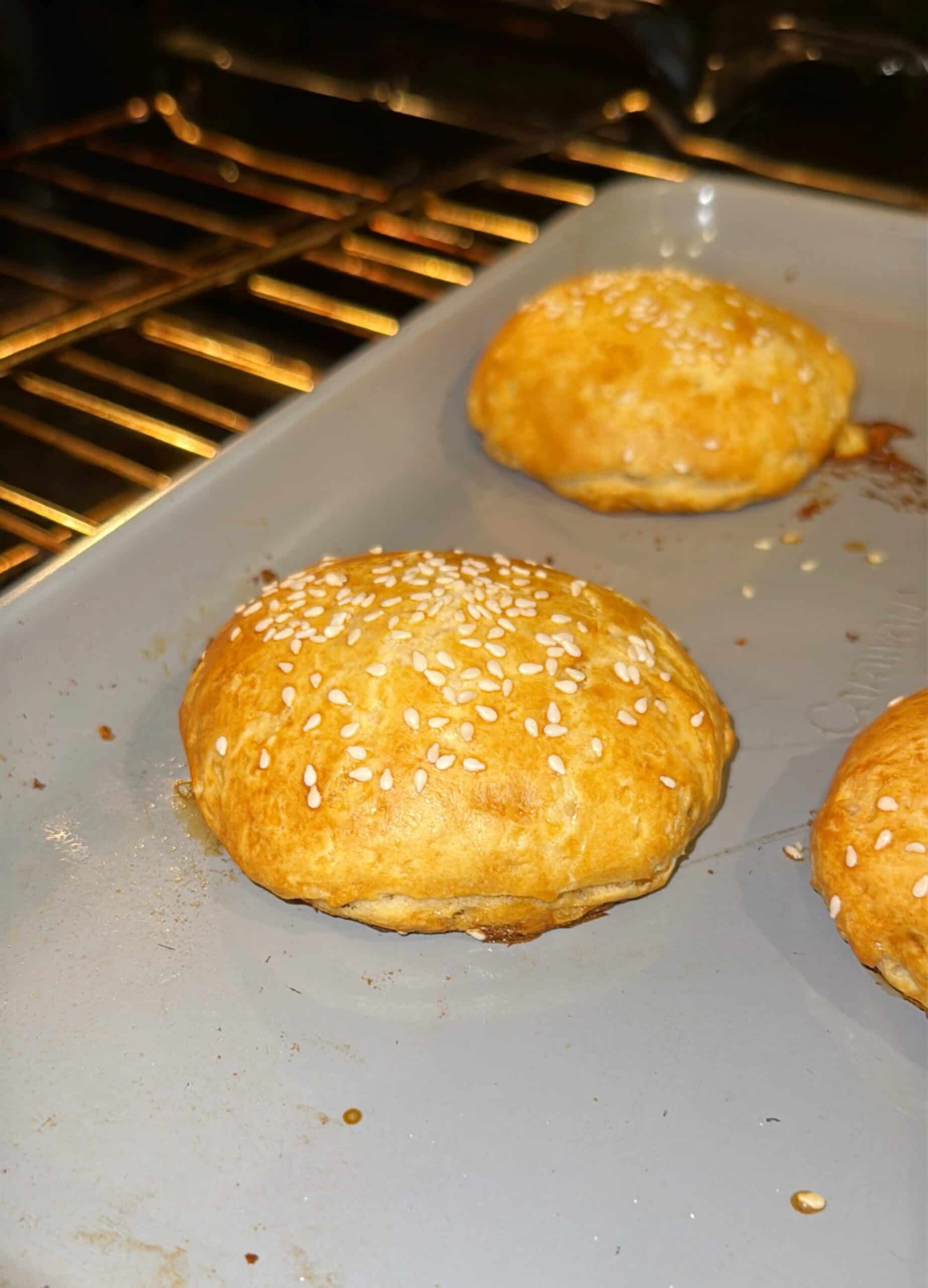 Golden, round pastries topped with sesame seeds are baking on a gray tray in an oven. The pastries appear puffy and slightly browned.