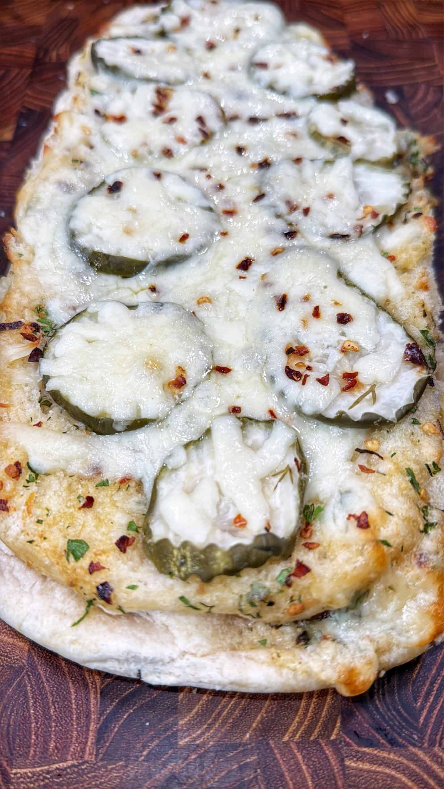A flatbread topped with melted cheese, pickle slices, herbs, and red pepper flakes, served on a wooden surface.