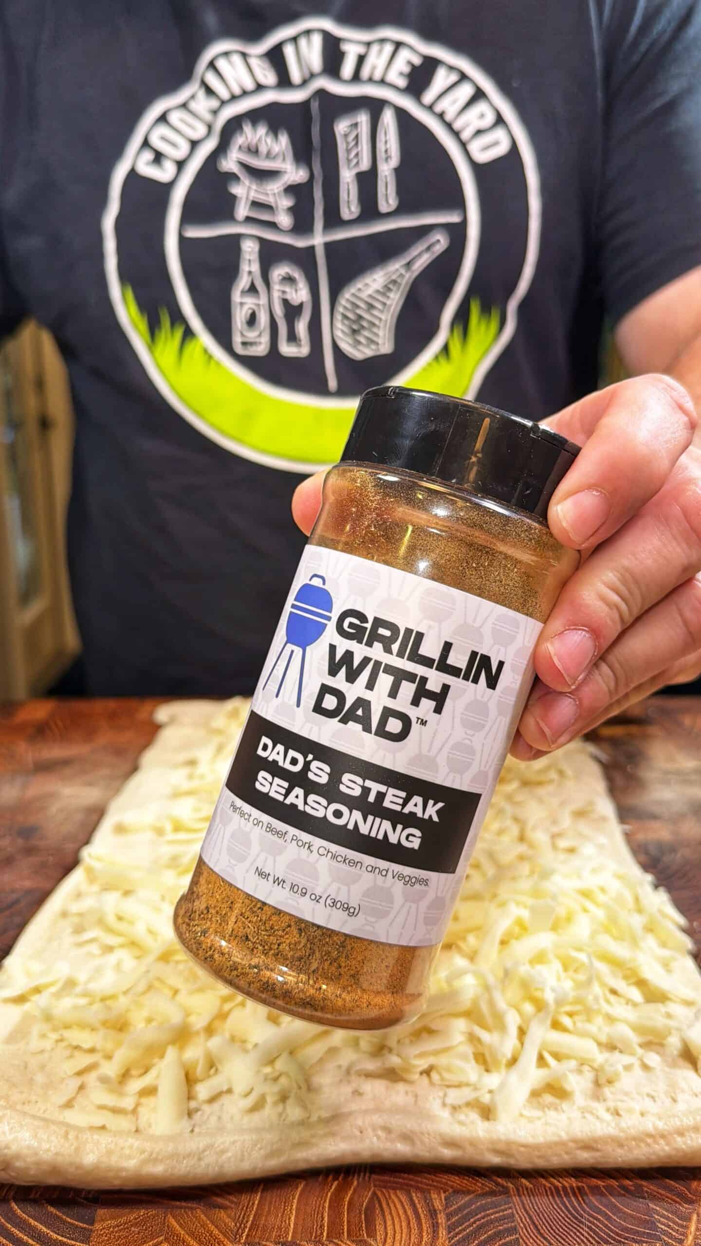A person wearing a Cookin in the Yard shirt sprinkles Grillin With Dad Dads Steak Seasoning over a sheet of dough topped with shredded cheese.