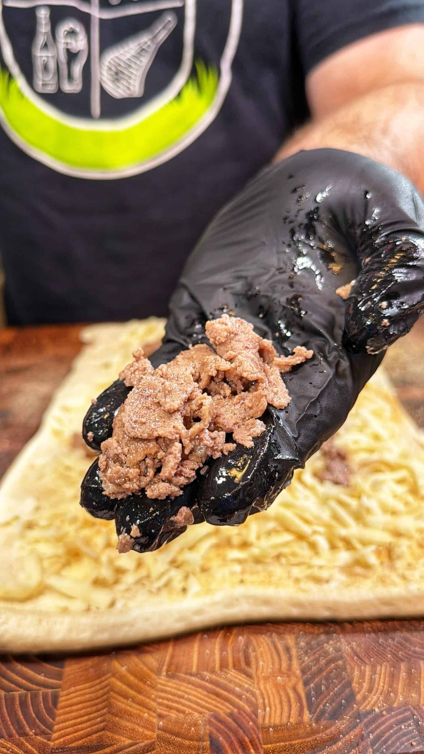 A person wearing a black glove holds a small portion of seasoned, cooked ground meat over a piece of flat dough on a wooden surface.