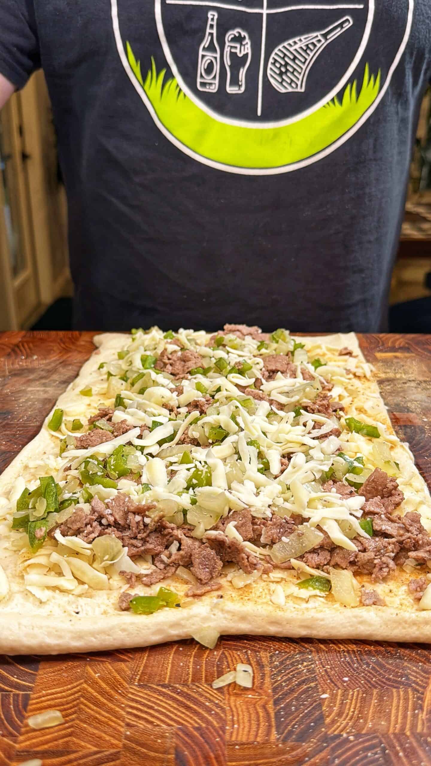 A person wearing a black T-shirt with food and drink graphics stands behind a wooden surface, preparing a flatbread topped with chopped meat, green peppers, onions, and shredded cheese.