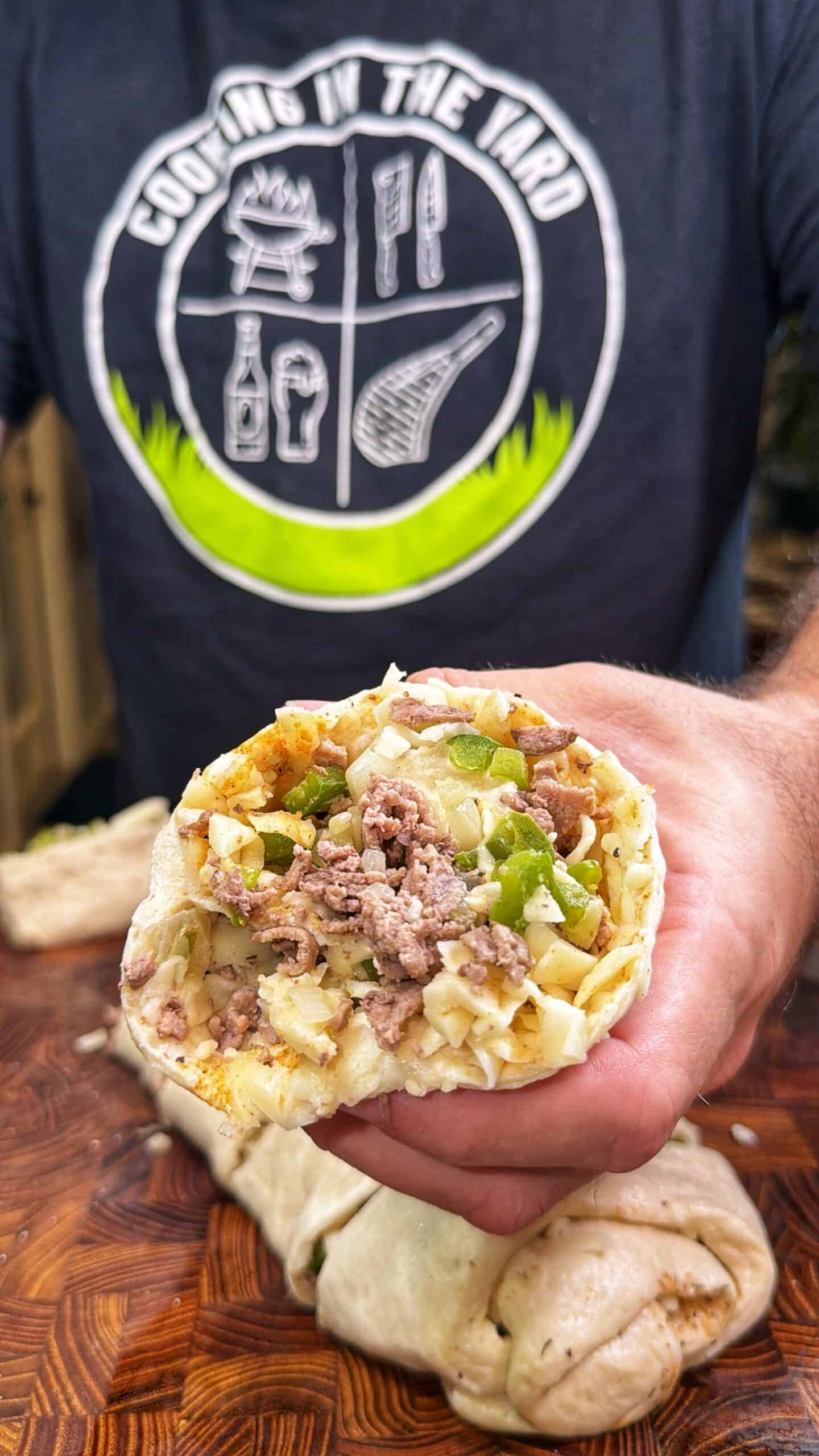 A person wearing a Cooking in the Yard shirt holds a burrito cut open, revealing ground beef, chopped onions, green peppers, and other ingredients on a wooden surface.